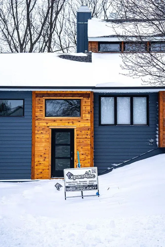 Modern home with wood accents and dark blue siding, covered in snow. A sign stands in the yard.