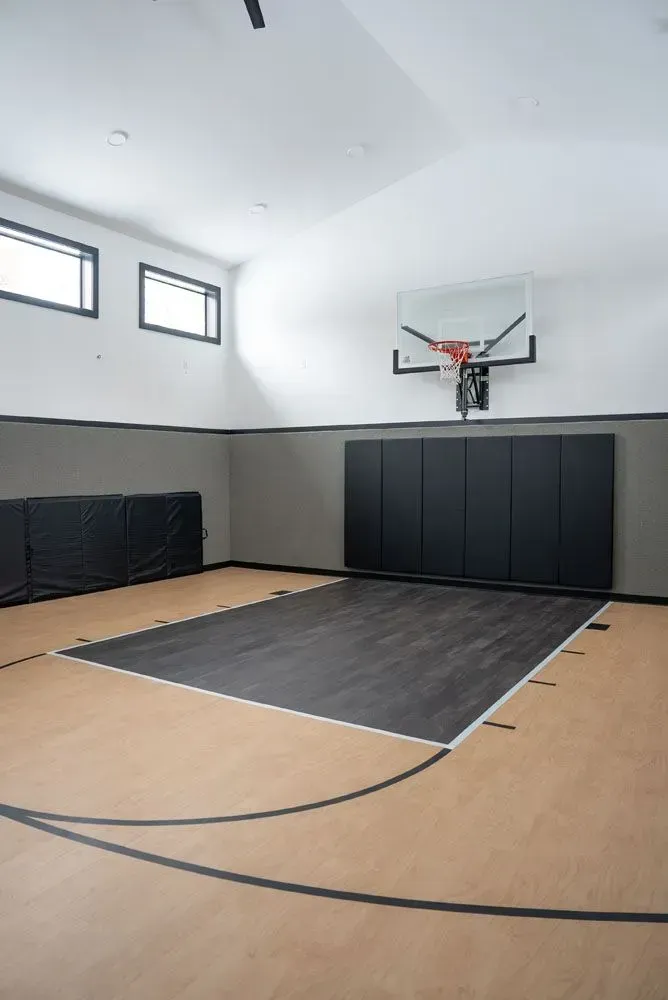 Indoor basketball court with black and tan flooring, black backboards, and a hoop.