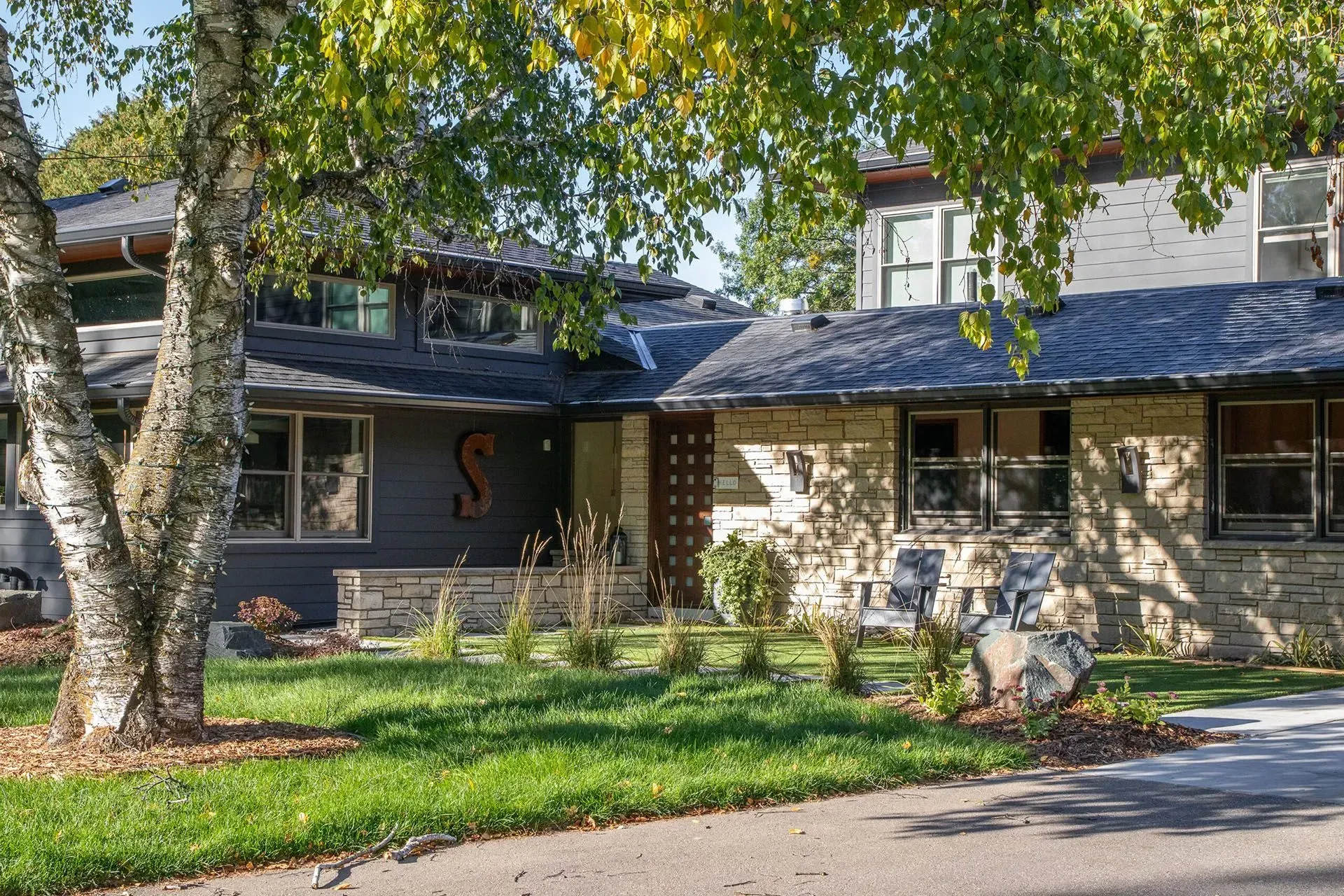 A house with stone and dark siding, a mature tree, and green grass.