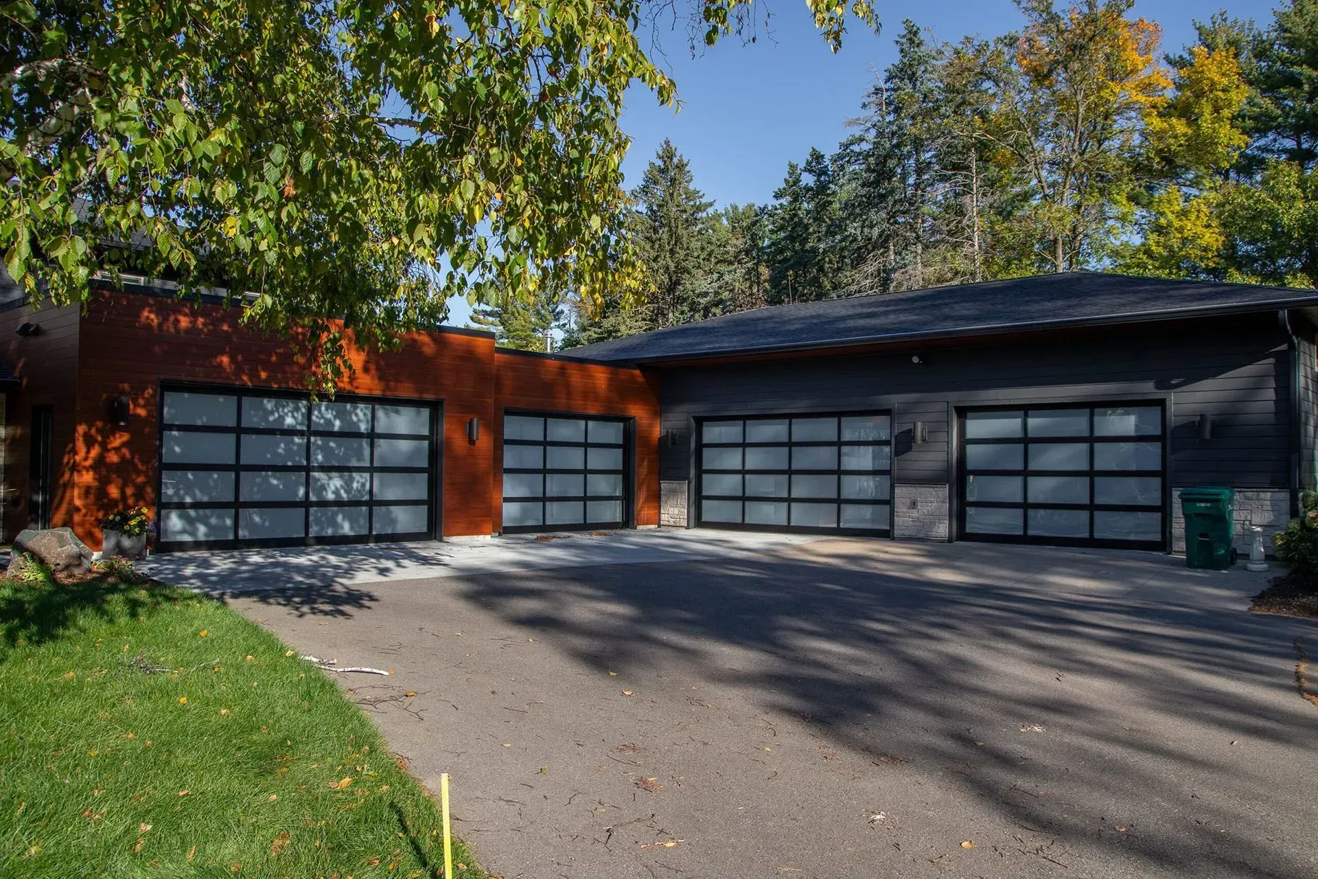 Garage with three glass-paneled doors, asphalt driveway, and green lawn. Exterior is brick and dark siding with trees in background.