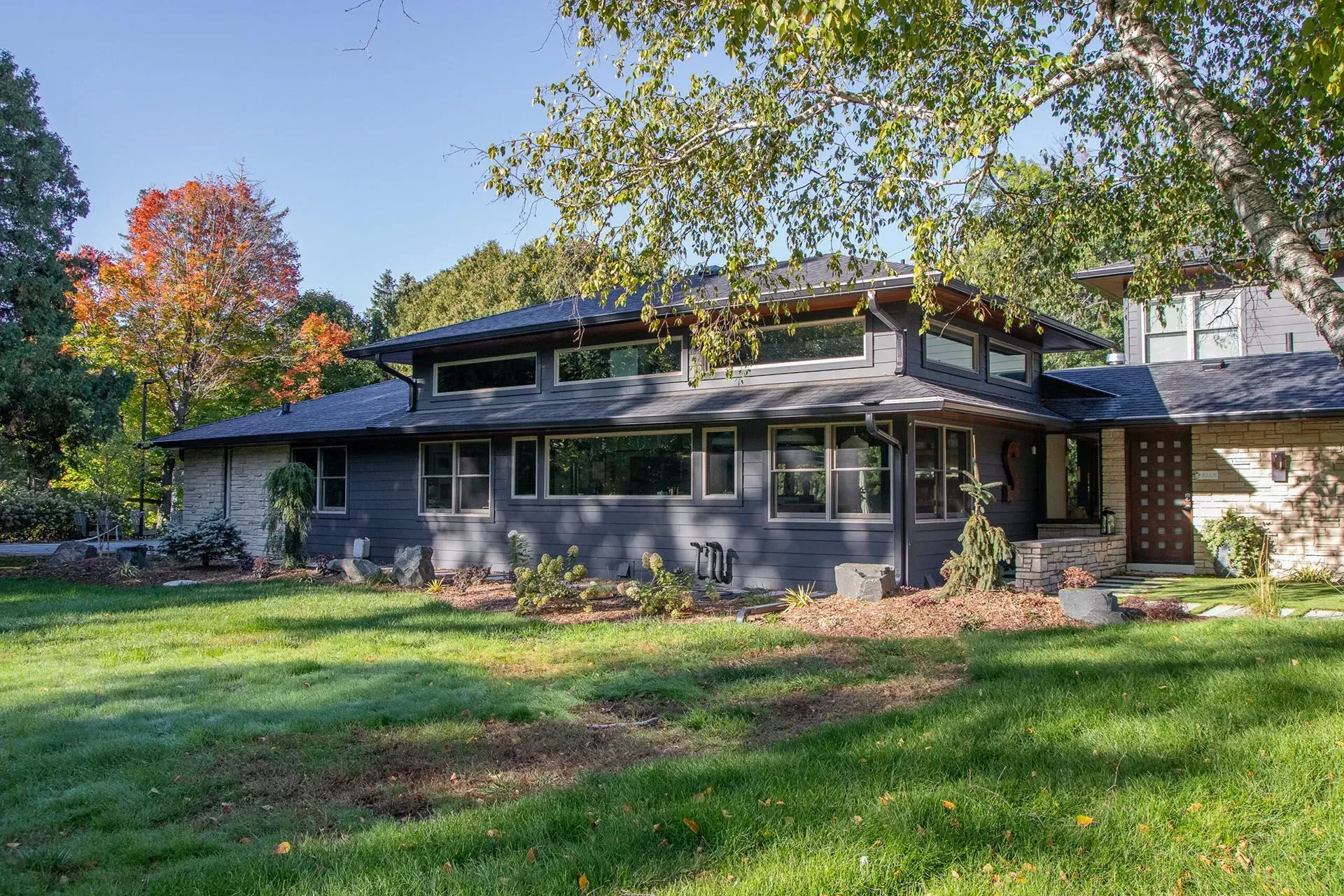 Gray house with dark roof and multiple windows, surrounded by green lawn and autumn trees.
