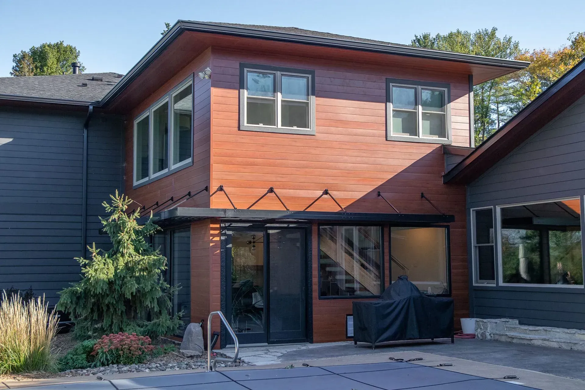 Two-story house addition with brown siding, large windows, and an enclosed patio by a pool.