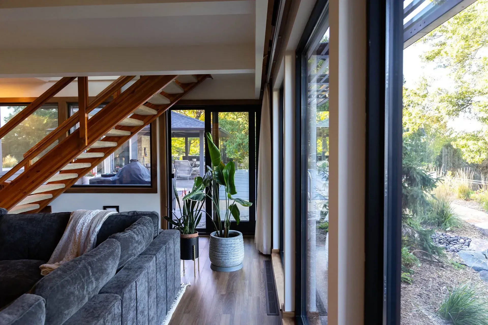 Living room with a large gray sofa, wood stairs, and floor-to-ceiling windows overlooking greenery.