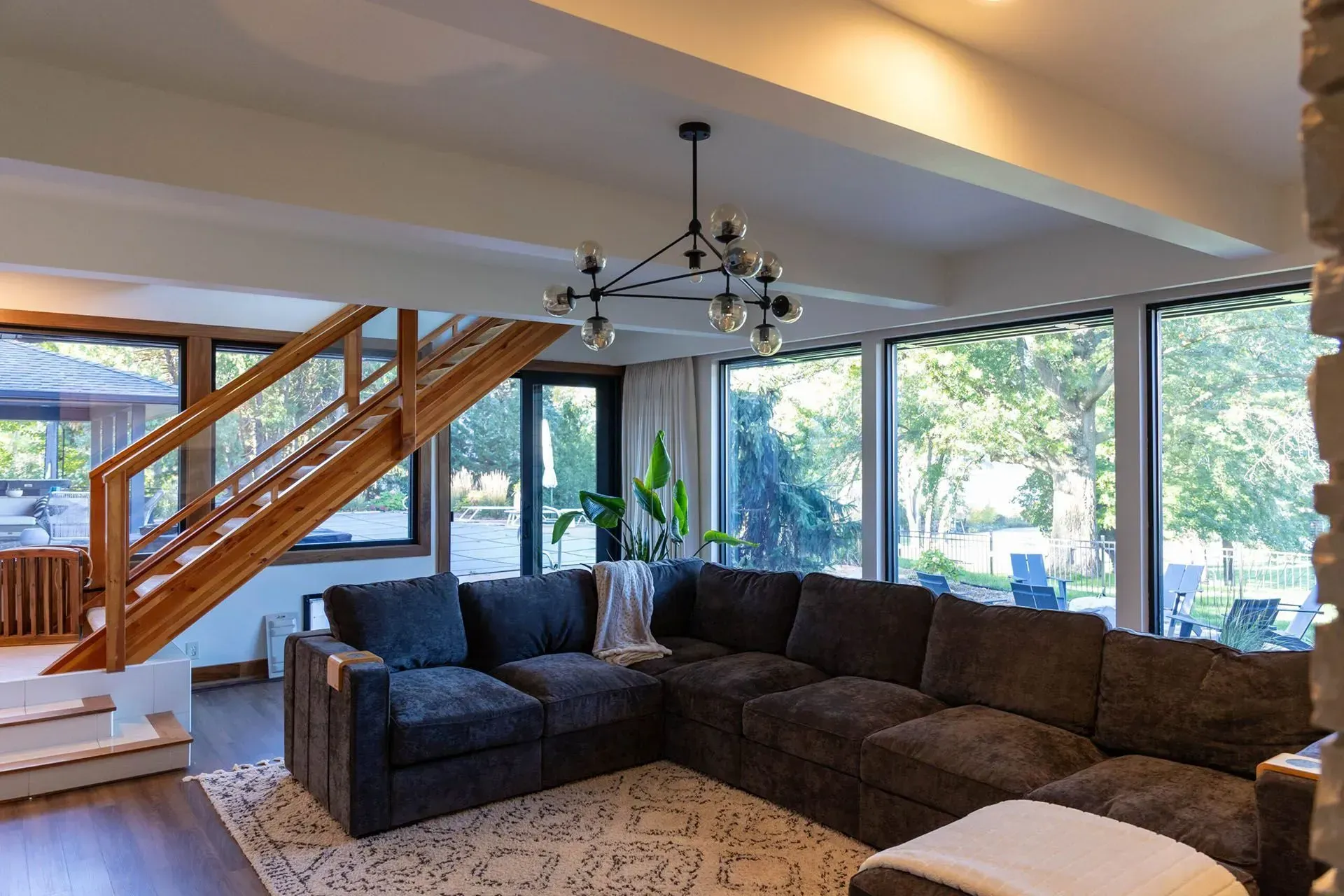 Living room with dark gray sectional sofa, large windows, wooden staircase, and modern chandelier.