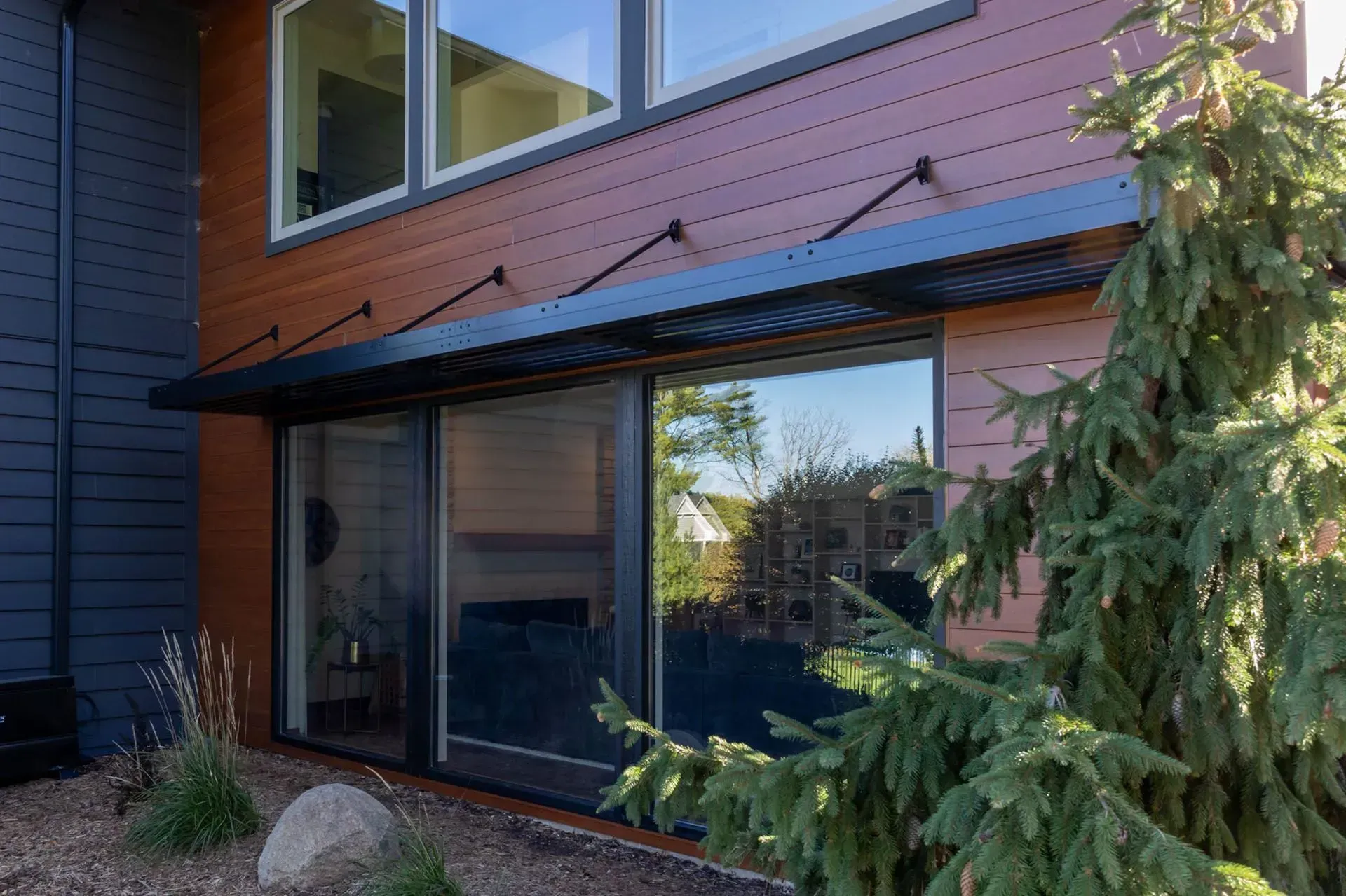 Modern building exterior with brown siding, large windows, black awning, and a small evergreen tree.