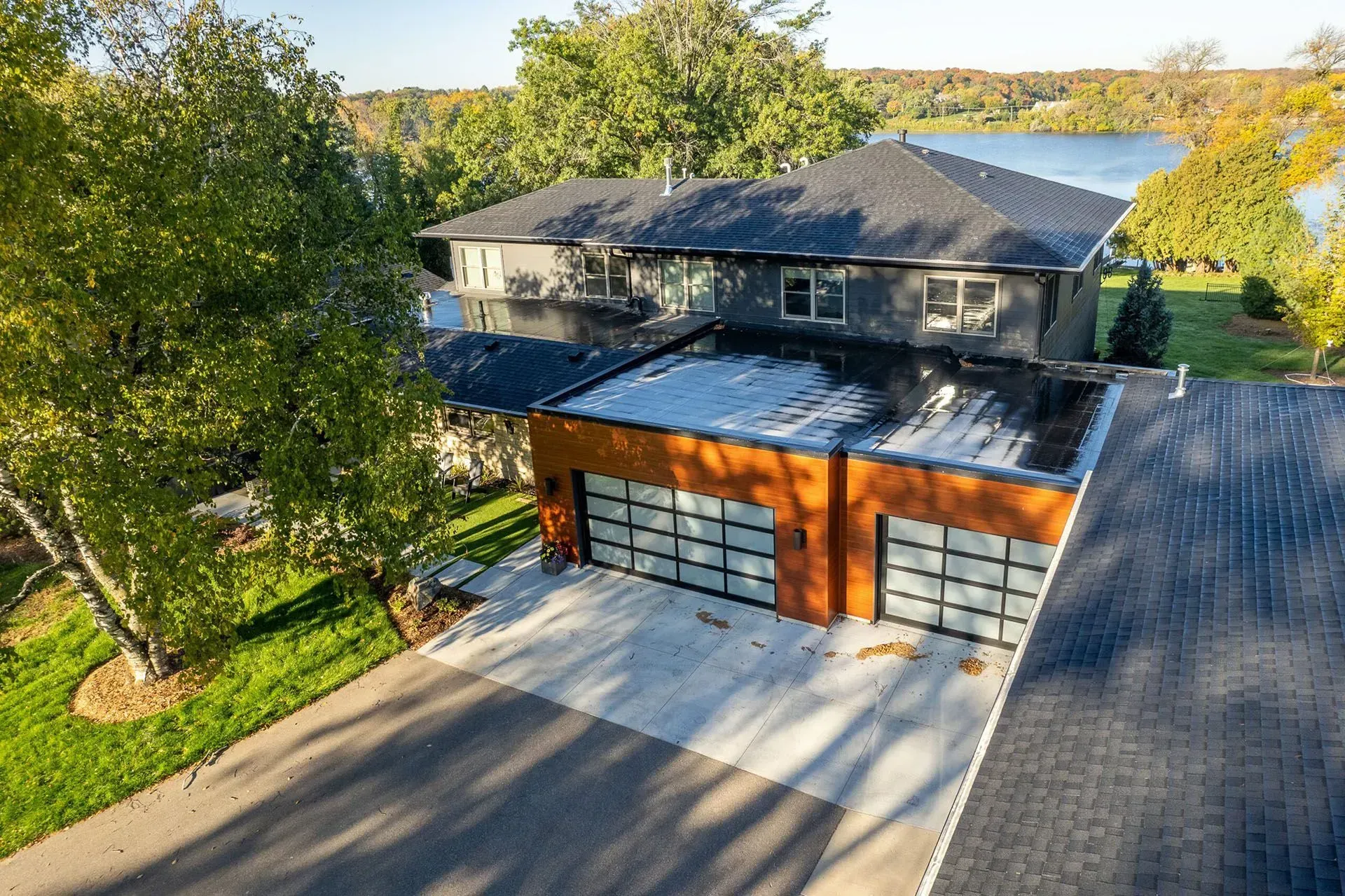 Modern two-story house with a flat-roof garage, glass doors, and a lake view. Green lawn and trees surround the home.
