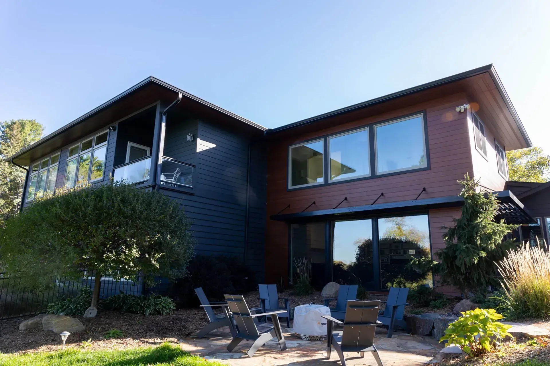 Modern home exterior with large windows, fire pit, and blue siding. Brown trim and a bright sunny day.