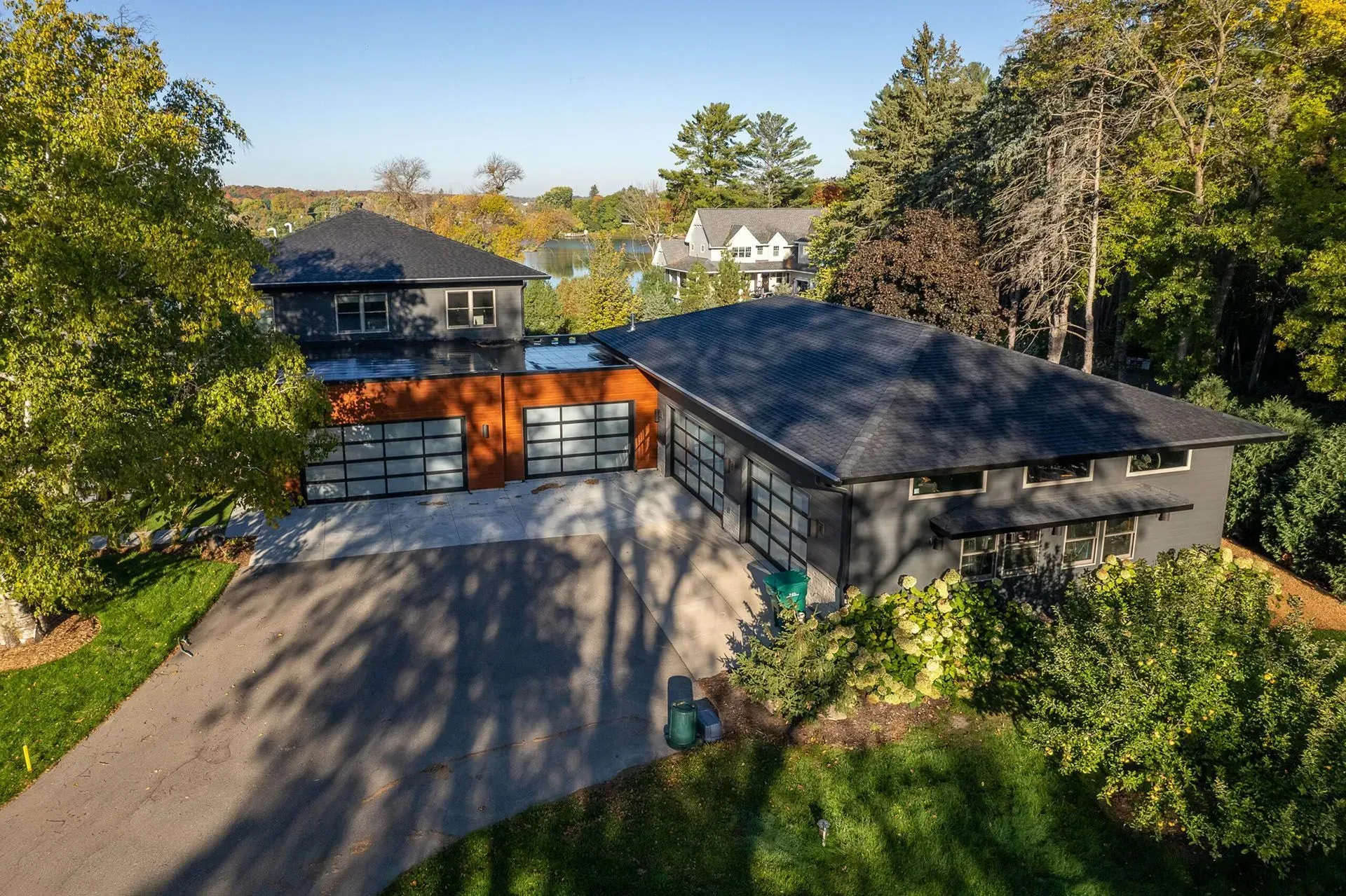 Modern gray house with black roof, orange garage doors, and a long driveway surrounded by green trees.