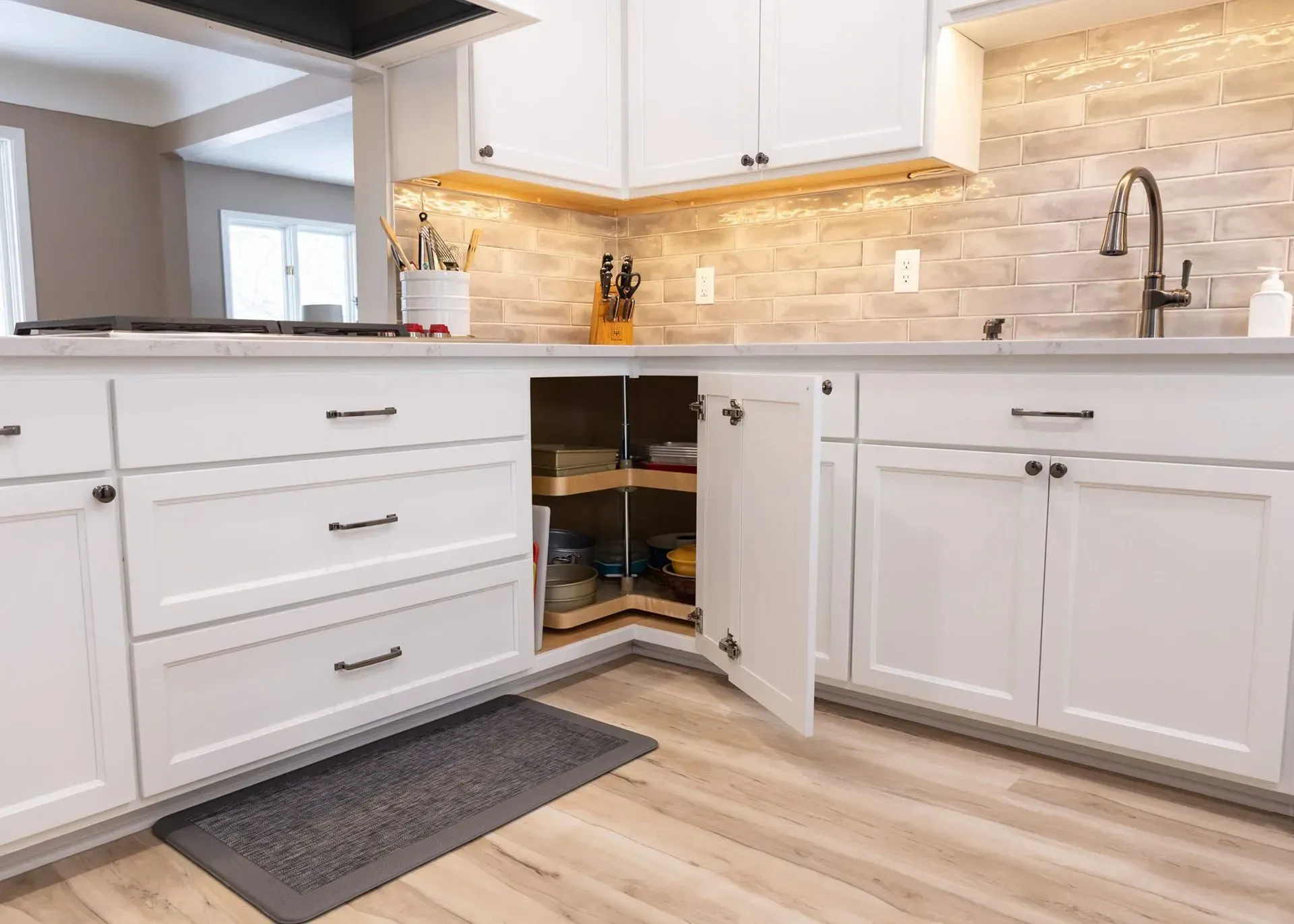 White kitchen cabinets with an open corner cabinet revealing a rotating shelf, light wooden floor.