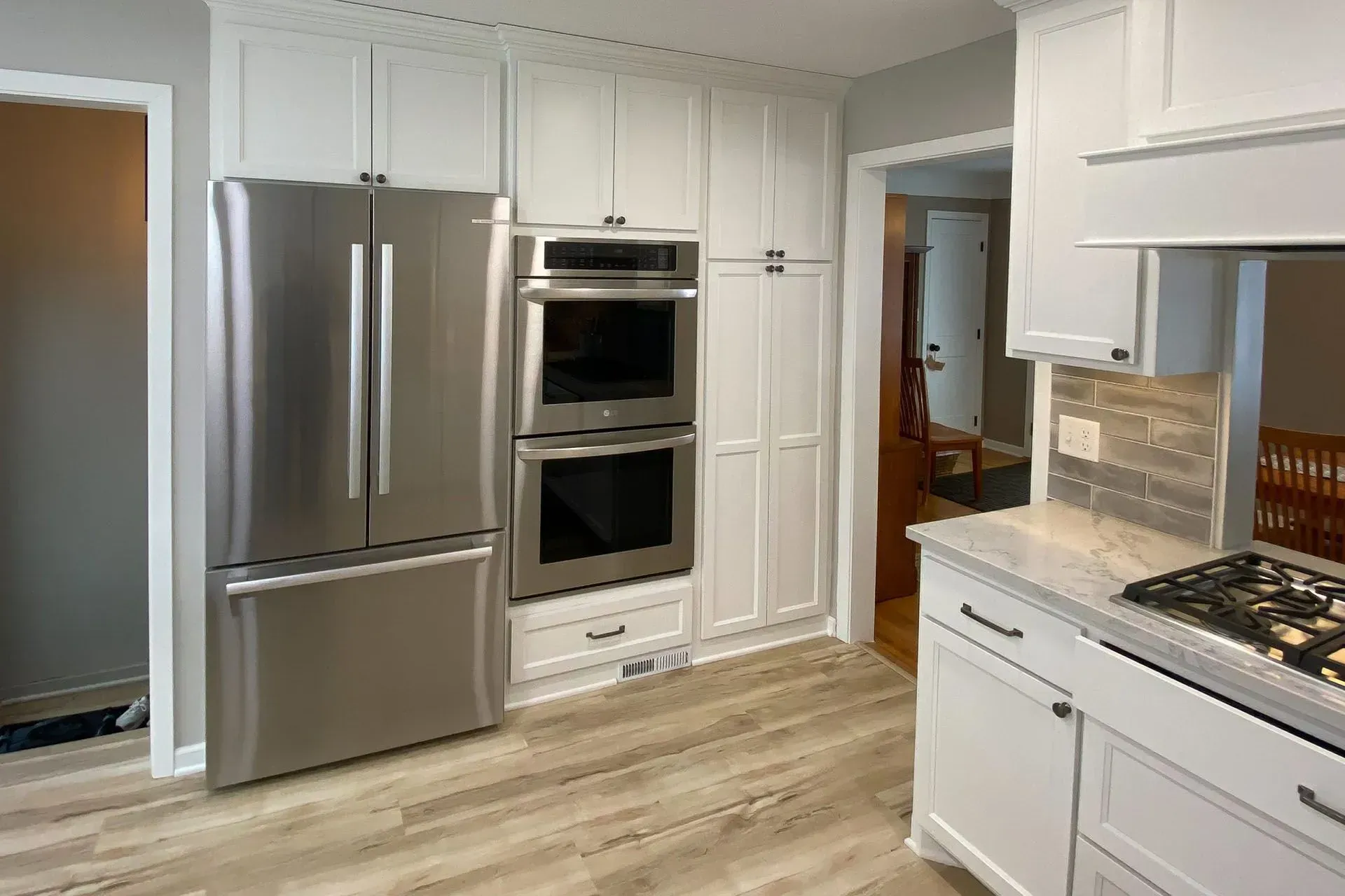White kitchen with stainless steel appliances: refrigerator, ovens, and cabinets. Light wood-look flooring.