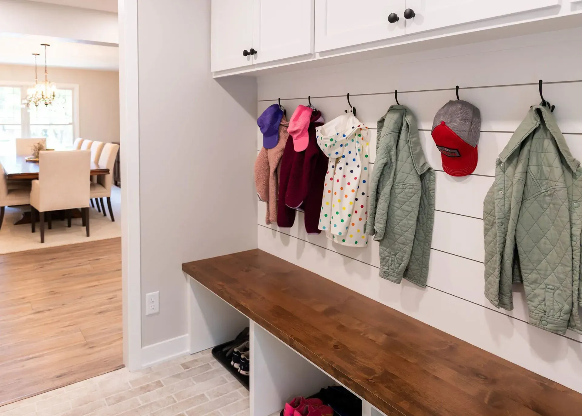 Mudroom with a wooden bench, hooks with coats, and cabinets. Dining room visible in the background.