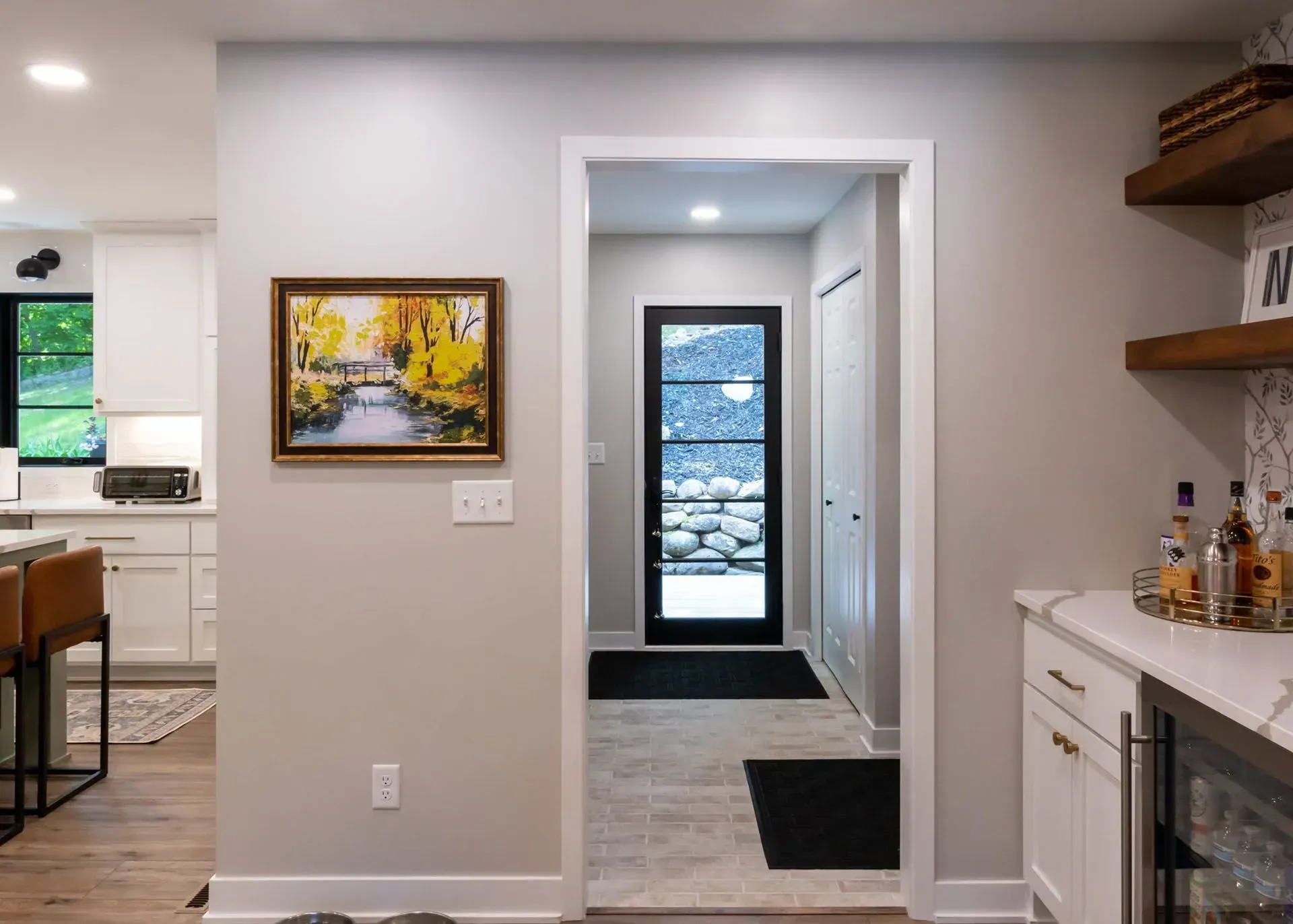 Interior view with doorway, artwork, kitchen, and bar area. Neutral walls, dark door, black rugs.