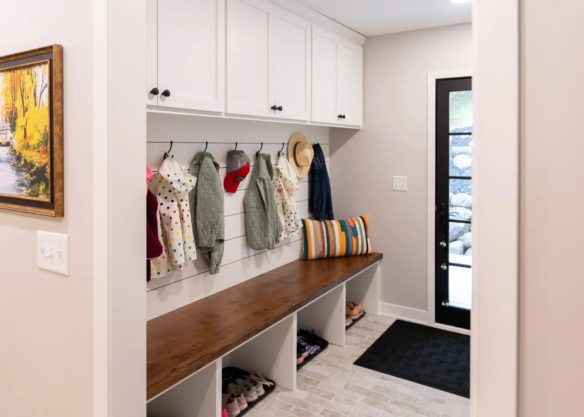 Mudroom with white cabinets, wooden bench, and coats hanging, leading to a black door.