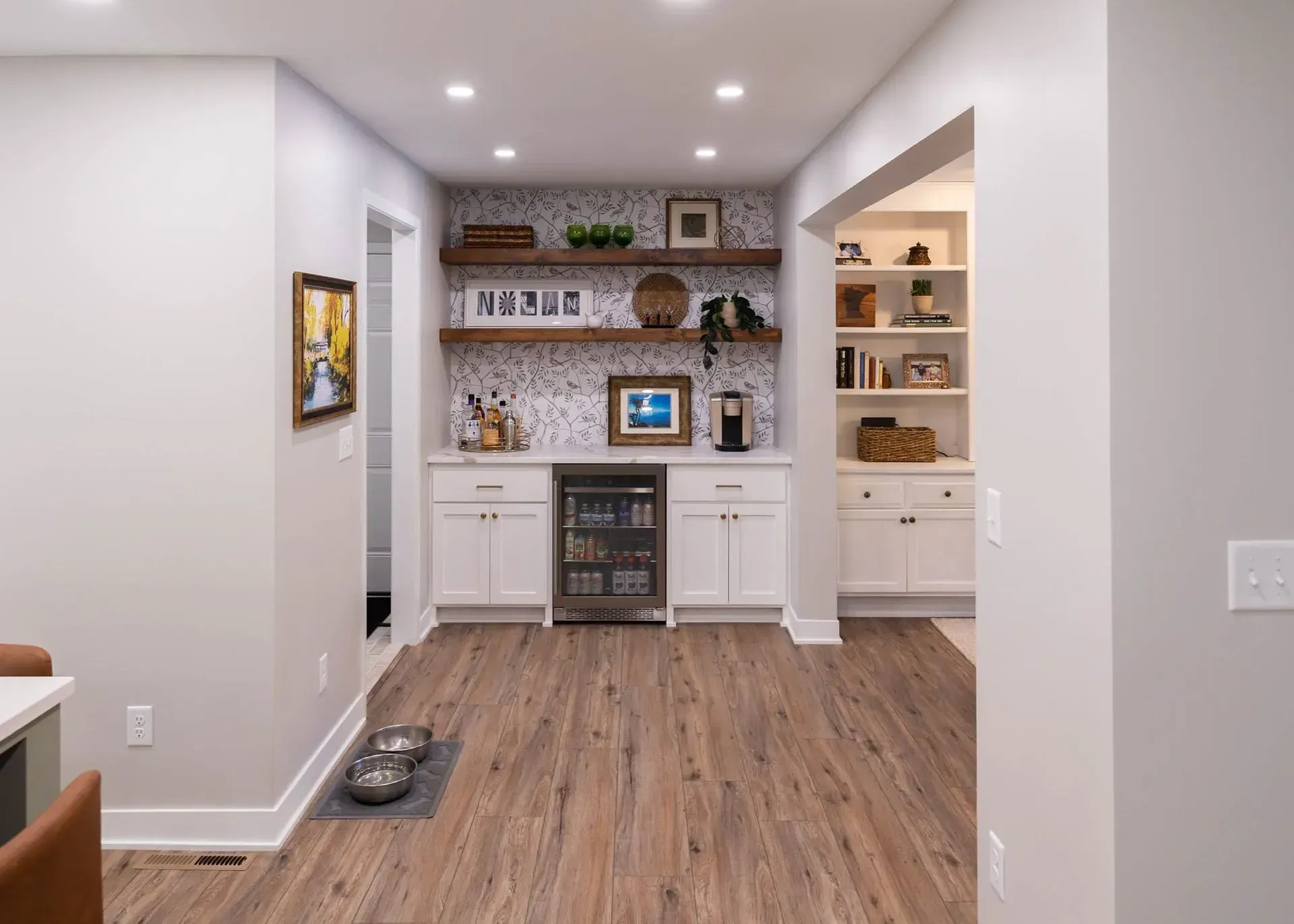 Modern built-in bar area with white cabinets, wood shelves, patterned wallpaper, and wood-look flooring.