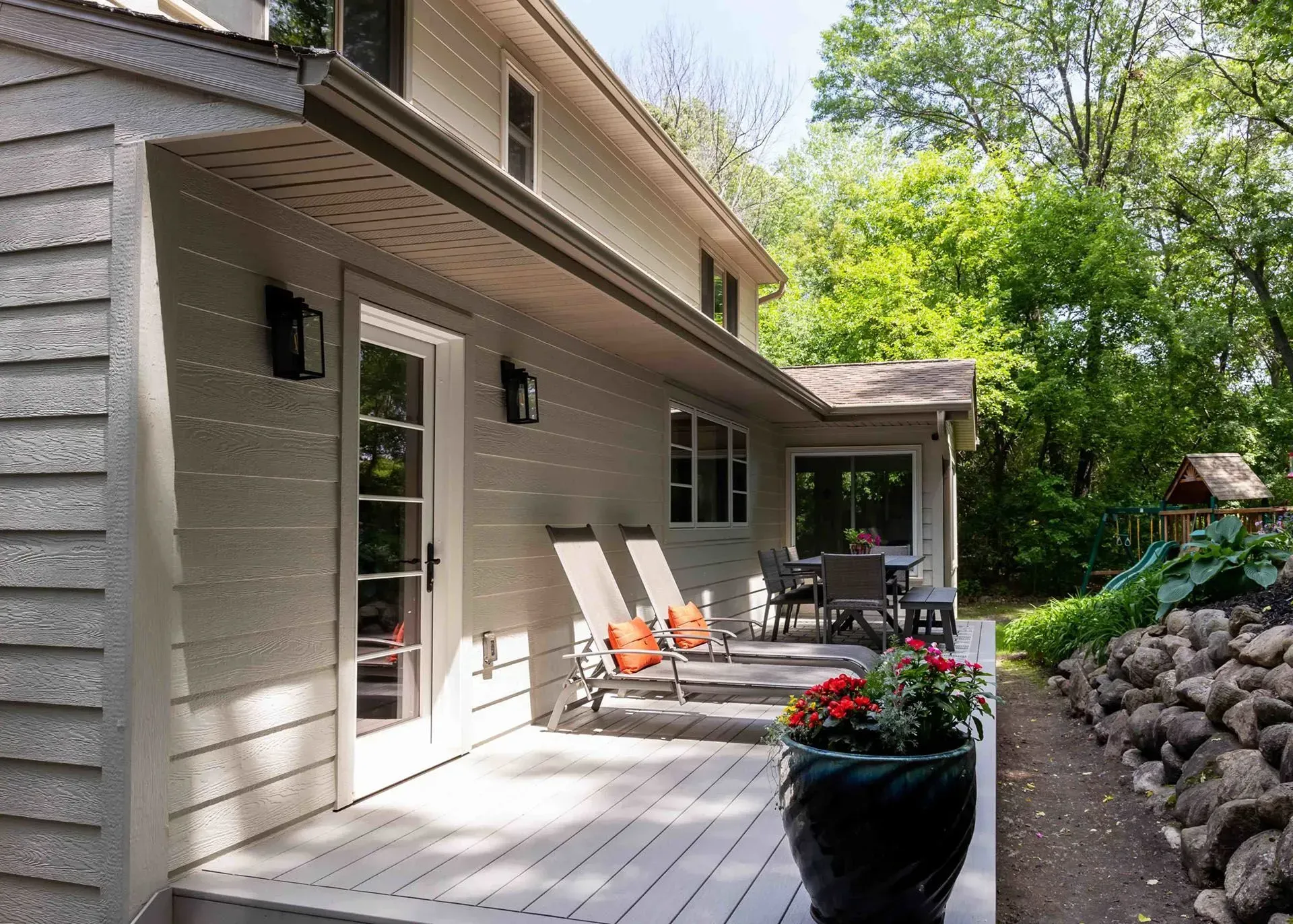 Back exterior of a two-story house with a wooden deck and patio furniture, surrounded by trees.