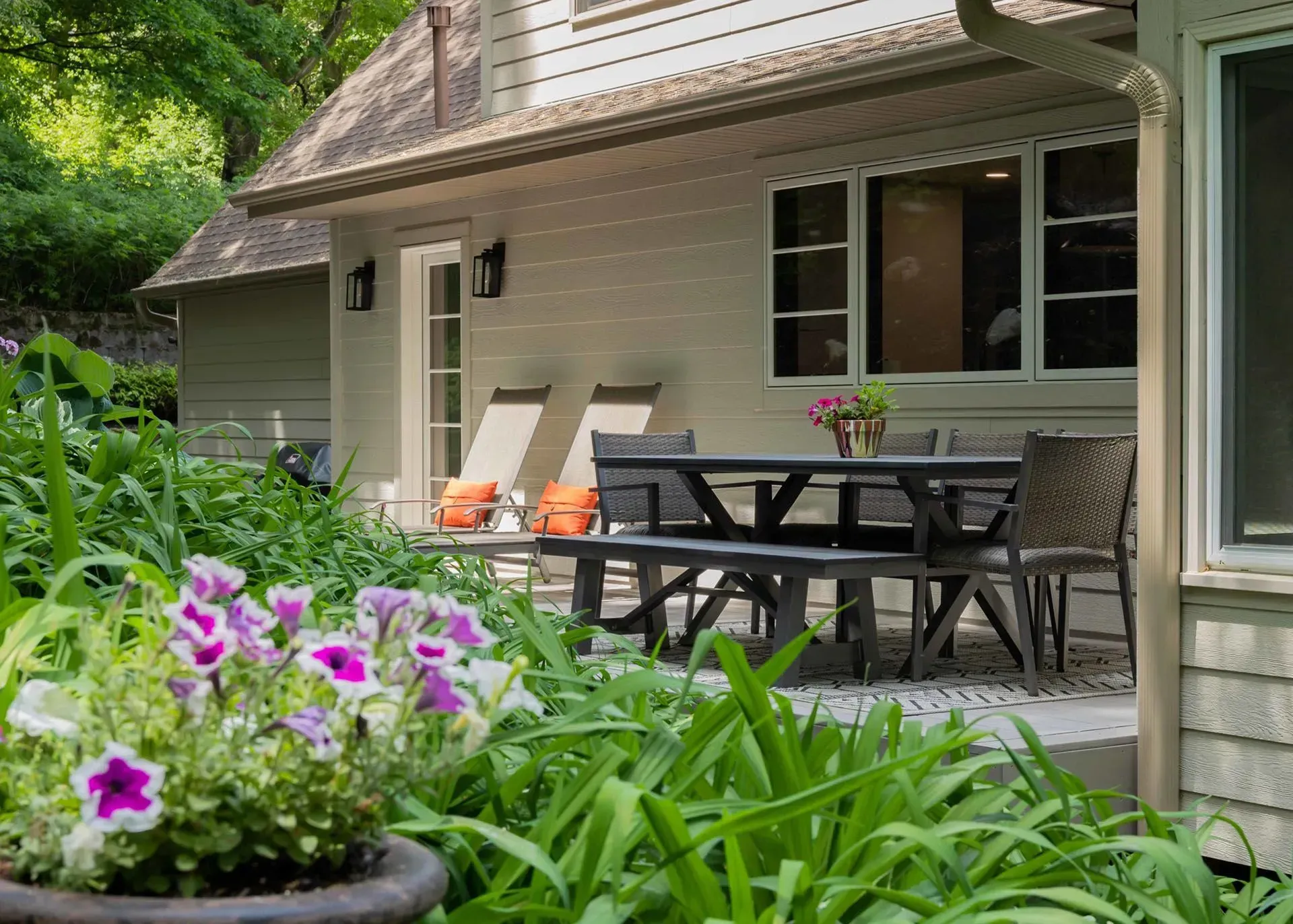 Outdoor patio with dark table and chairs, near a house with landscaping and flower pots.