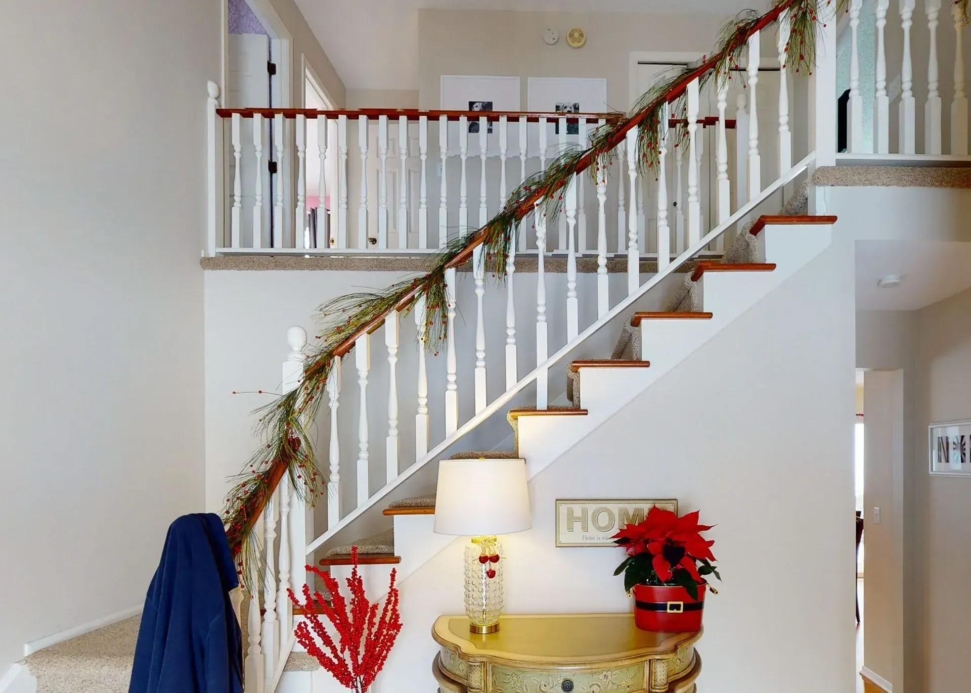 Staircase decorated for the holidays; garland, poinsettia, coat, and a table with a lamp are present.