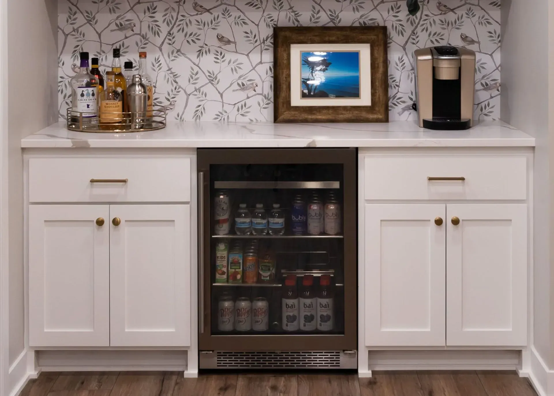 Built-in home bar with white cabinets, beverage fridge, and coffee maker. Liquor bottles and framed art sit on the countertop.