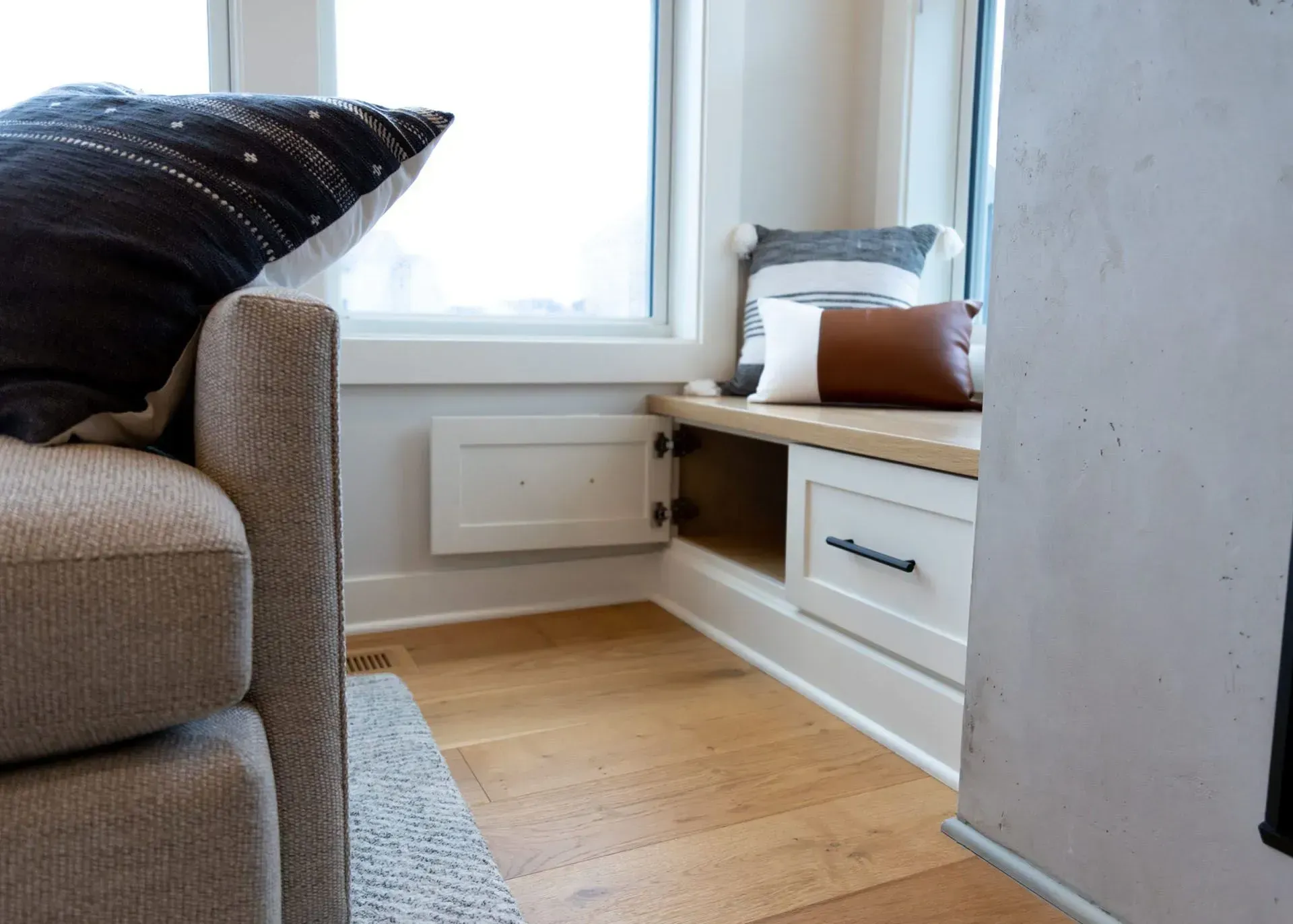 Cozy window seat with pillows. Beige sofa, white trim, wood floors, and a light-filled space.