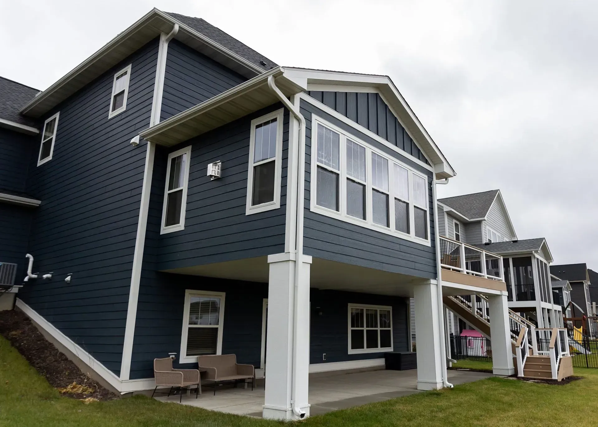 Blue-sided house with white trim, patio, and several windows on a cloudy day.
