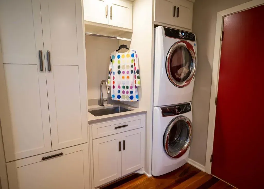 Laundry room with white cabinets, stacked washer/dryer, sink, and clothes hanging. Red door on the right.