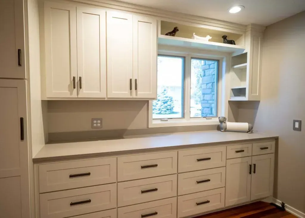 Cream-colored cabinetry with drawers, cabinets, and a counter surrounds a window, with black hardware.