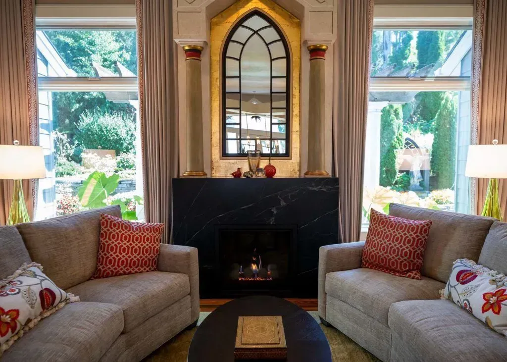 Living room with symmetrical design: sofas, fireplace, arched mirror, windows with lush outdoor view.