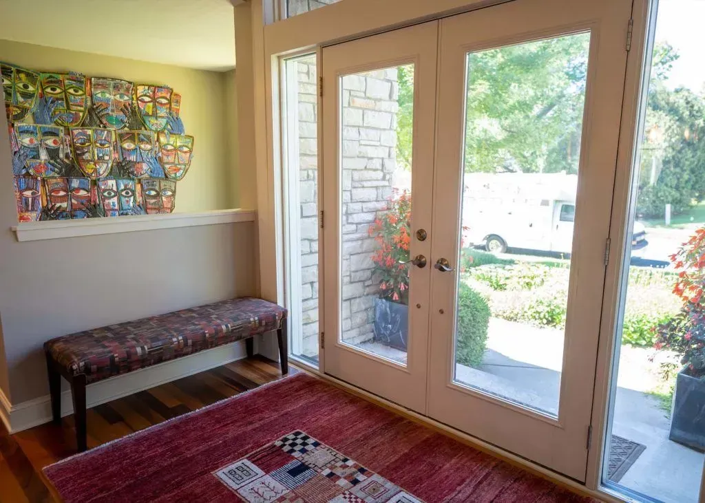 Entryway with double glass doors, bench, artwork, and outdoor view. Red rug on wood floor.