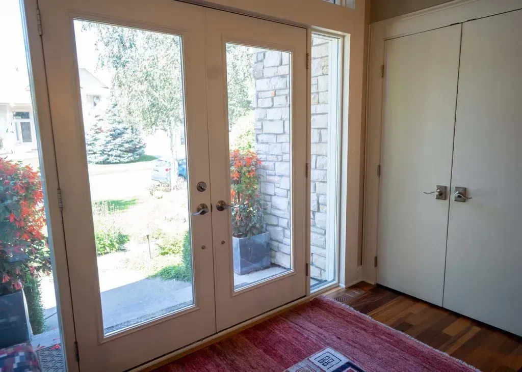 Double doors with sidelight, interior view. Red rug, light wood floor, white walls and trim, outside garden view.