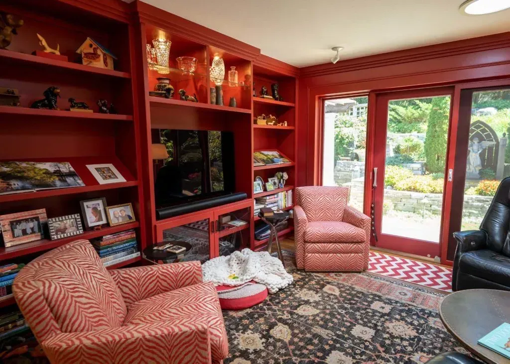 Red-walled living room with built-in shelves holding objects, TV, two patterned armchairs, and a view of a garden.