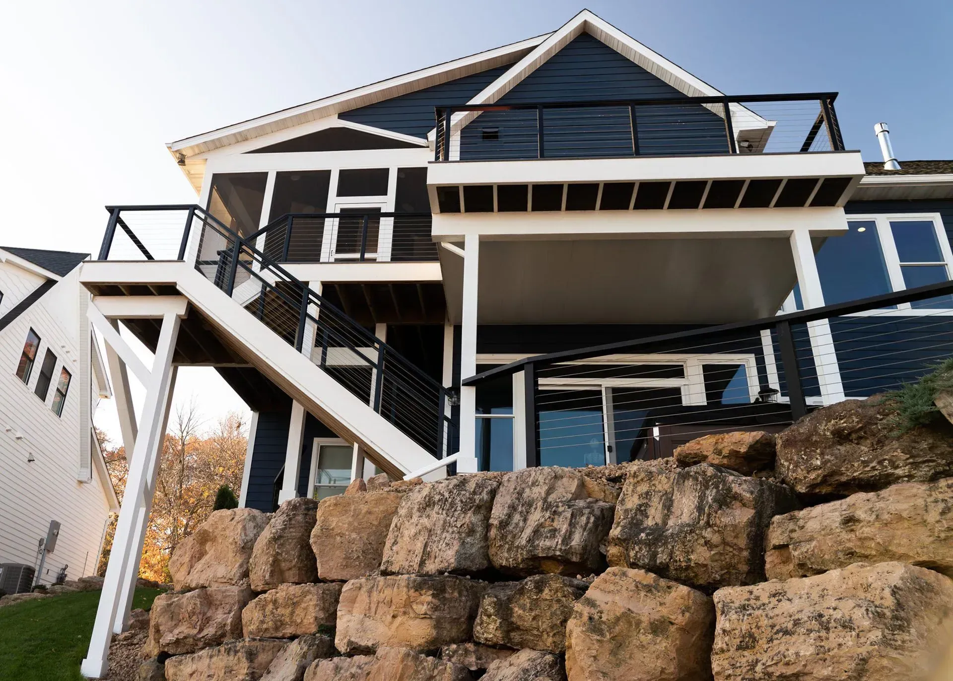 Dark blue house with multiple decks, stairs, and a rock retaining wall.