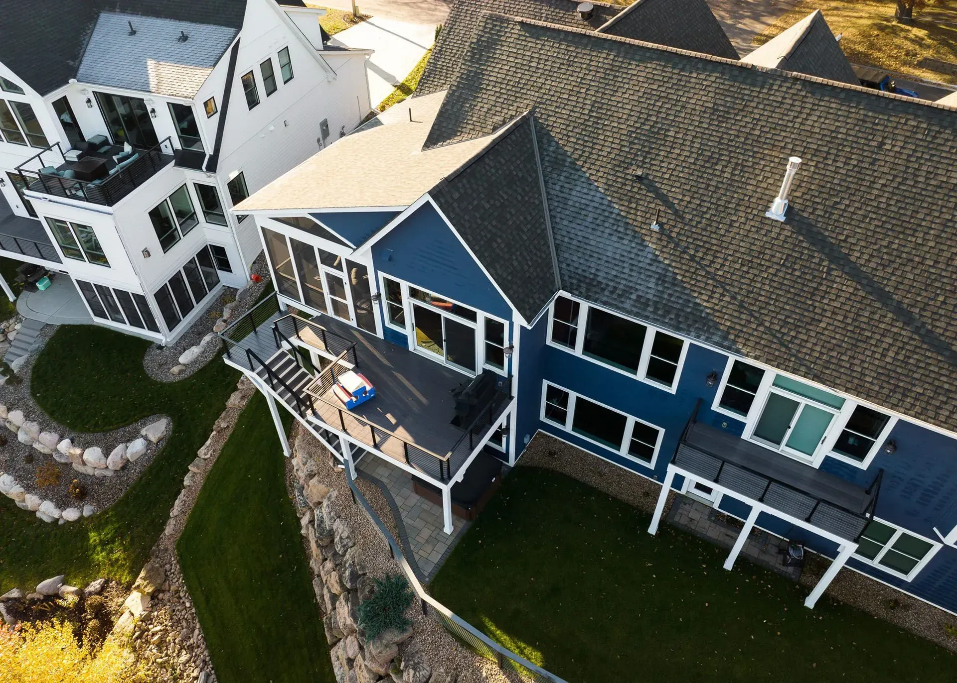Aerial view of a blue house with a deck and large windows, next to a white house with landscaping.