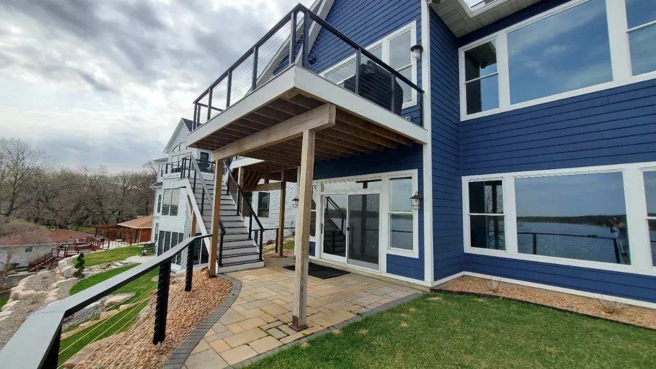 Blue house with decks, stairs, and glass railings, overlooking a lake, on a cloudy day.