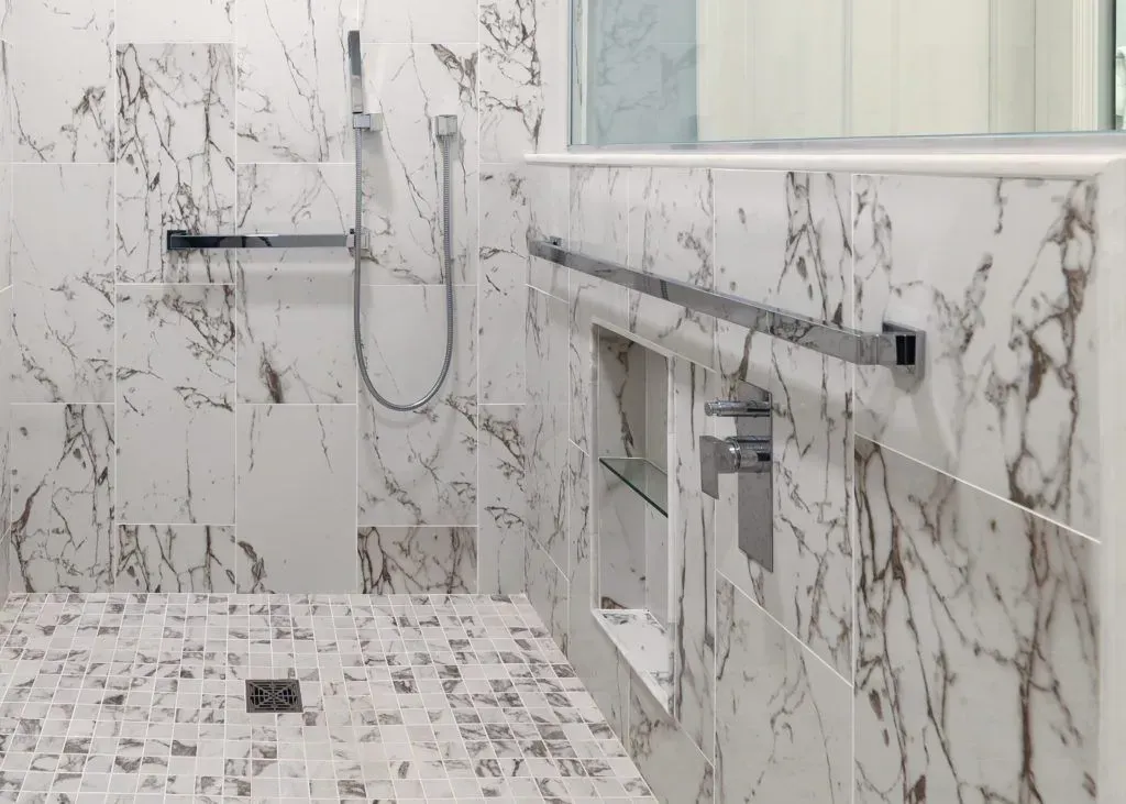 Marble-tiled shower with a hand-held showerhead and grab bars. Recessed shelf.