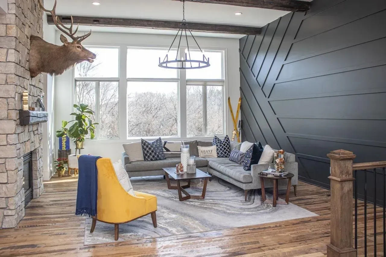 Living room with stone fireplace, elk head mount, couches, yellow chair, large windows, and dark accent wall.