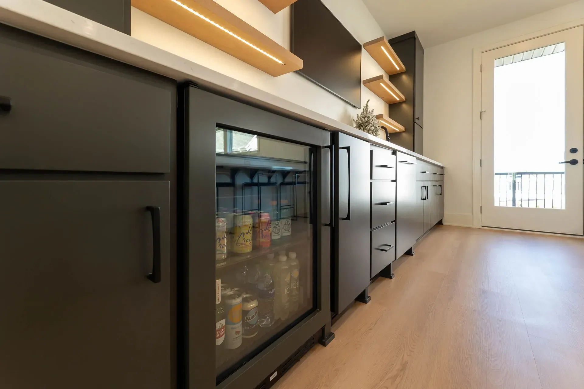 Dark cabinets with a glass-door refrigerator built-in, a light wood floor, and open shelves with lighting.