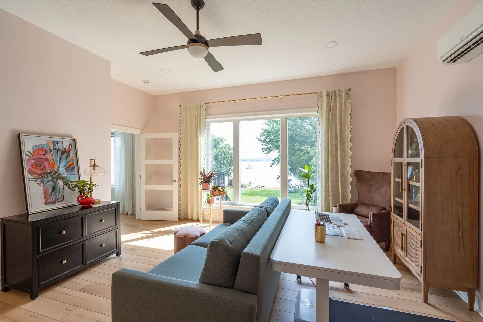 Living room with pink walls, sofa, table, cabinet, large window overlooking a yard and water.
