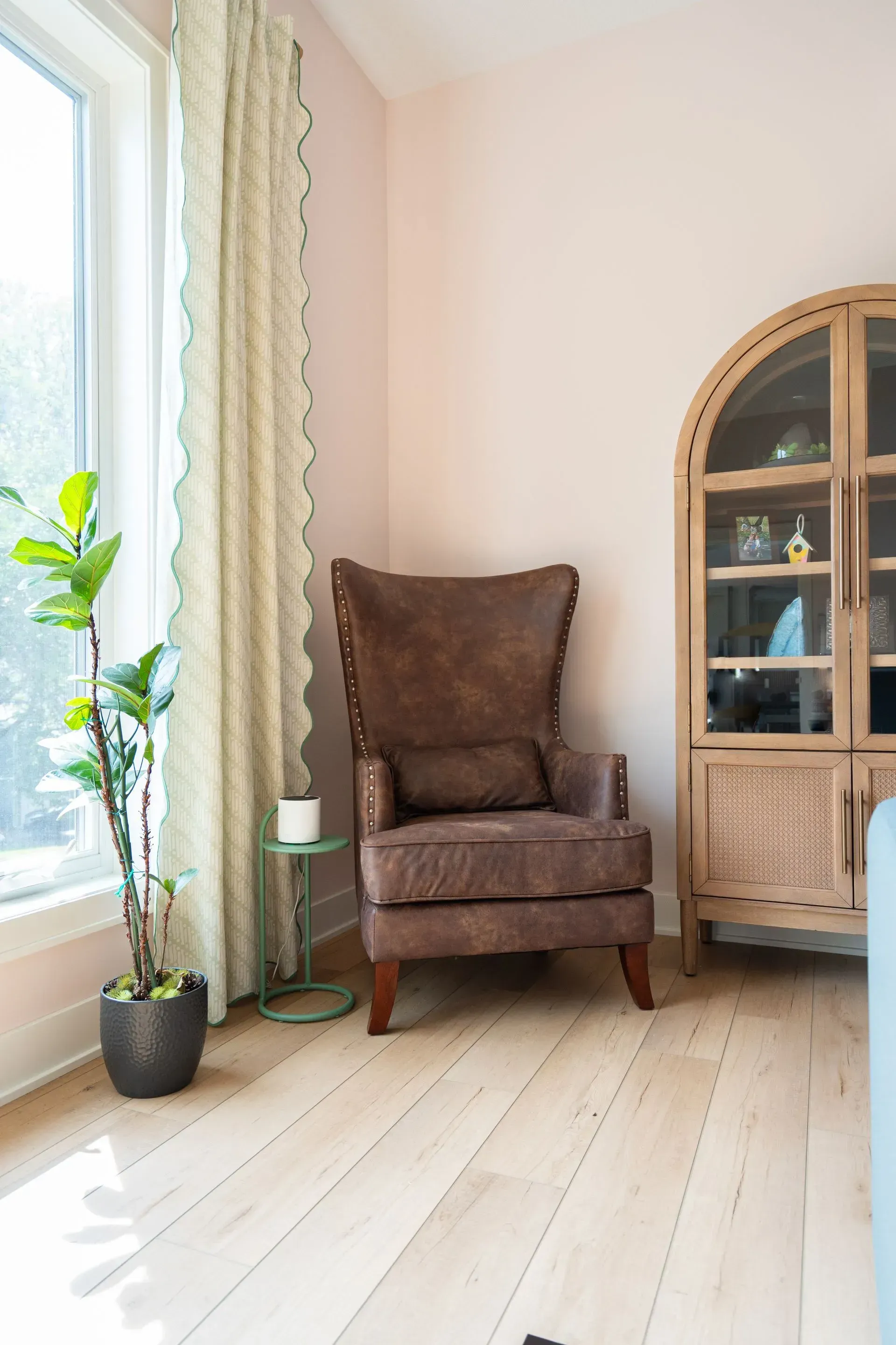 A brown armchair, a glass cabinet, and a potted plant in a room with light pink walls and light wood floors.