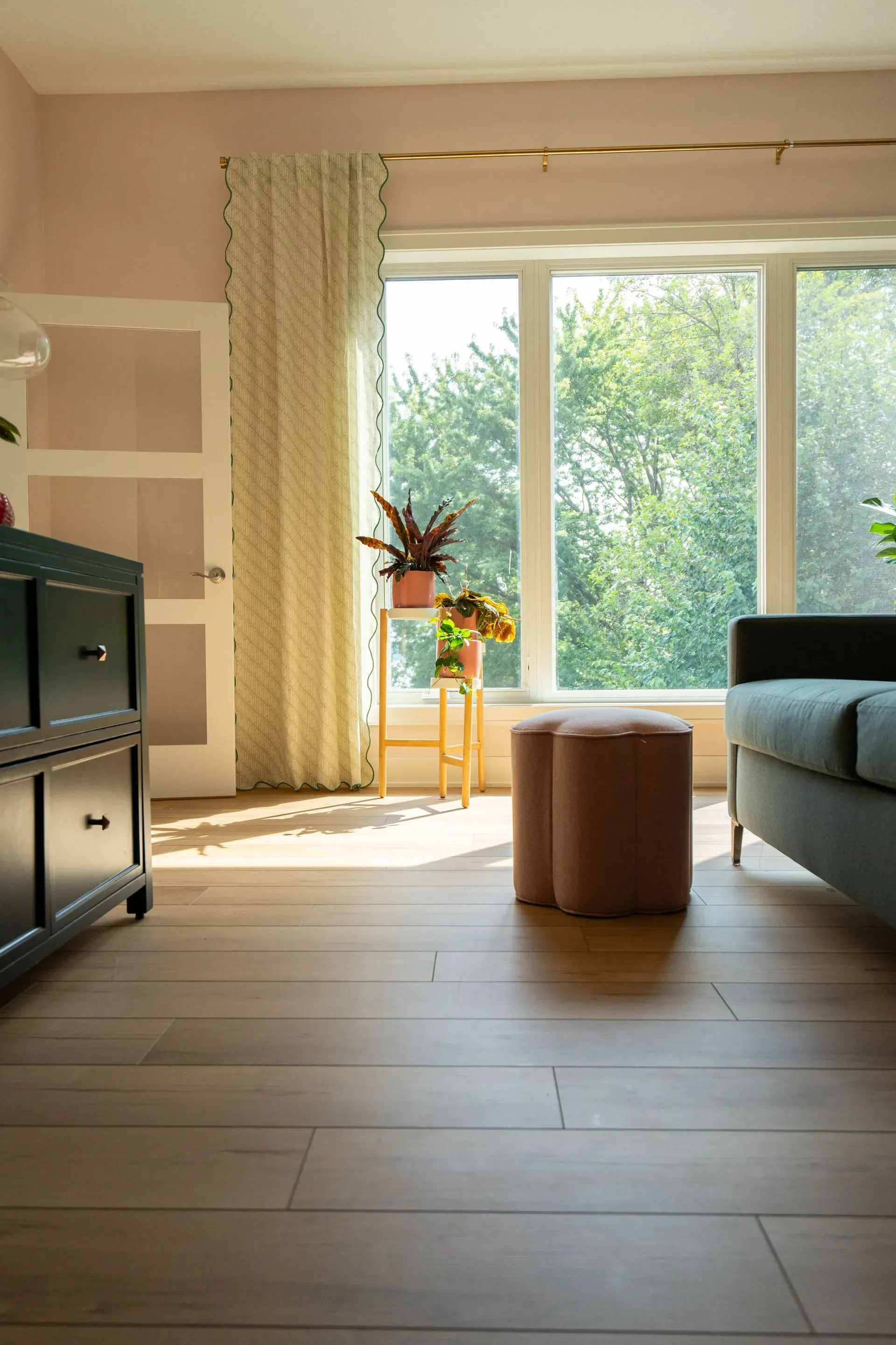 Living room with large window, potted plants, tan ottoman, and blue sofa.