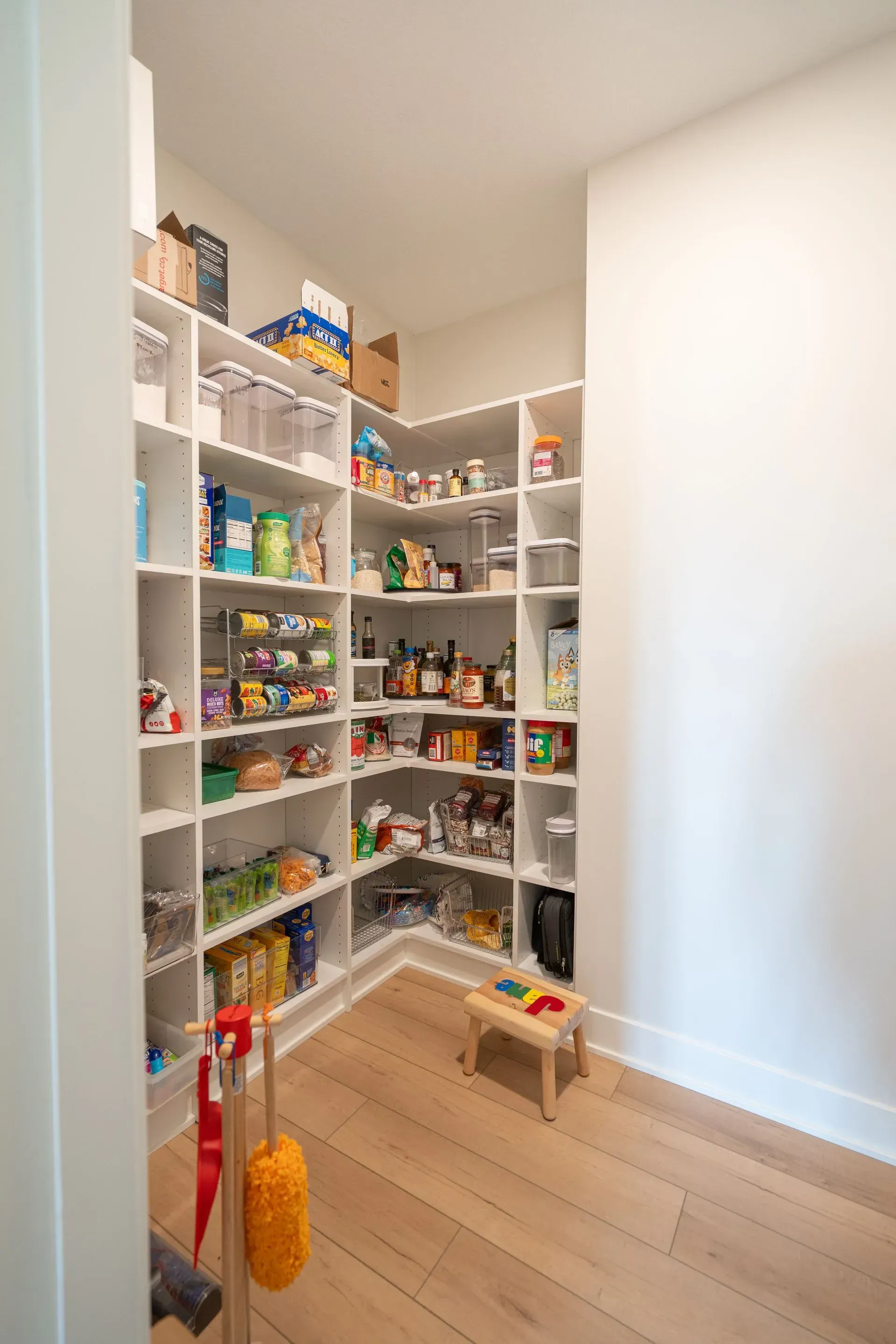 White, organized pantry with built-in shelves. Food items fill shelves, wooden floor, small stool.