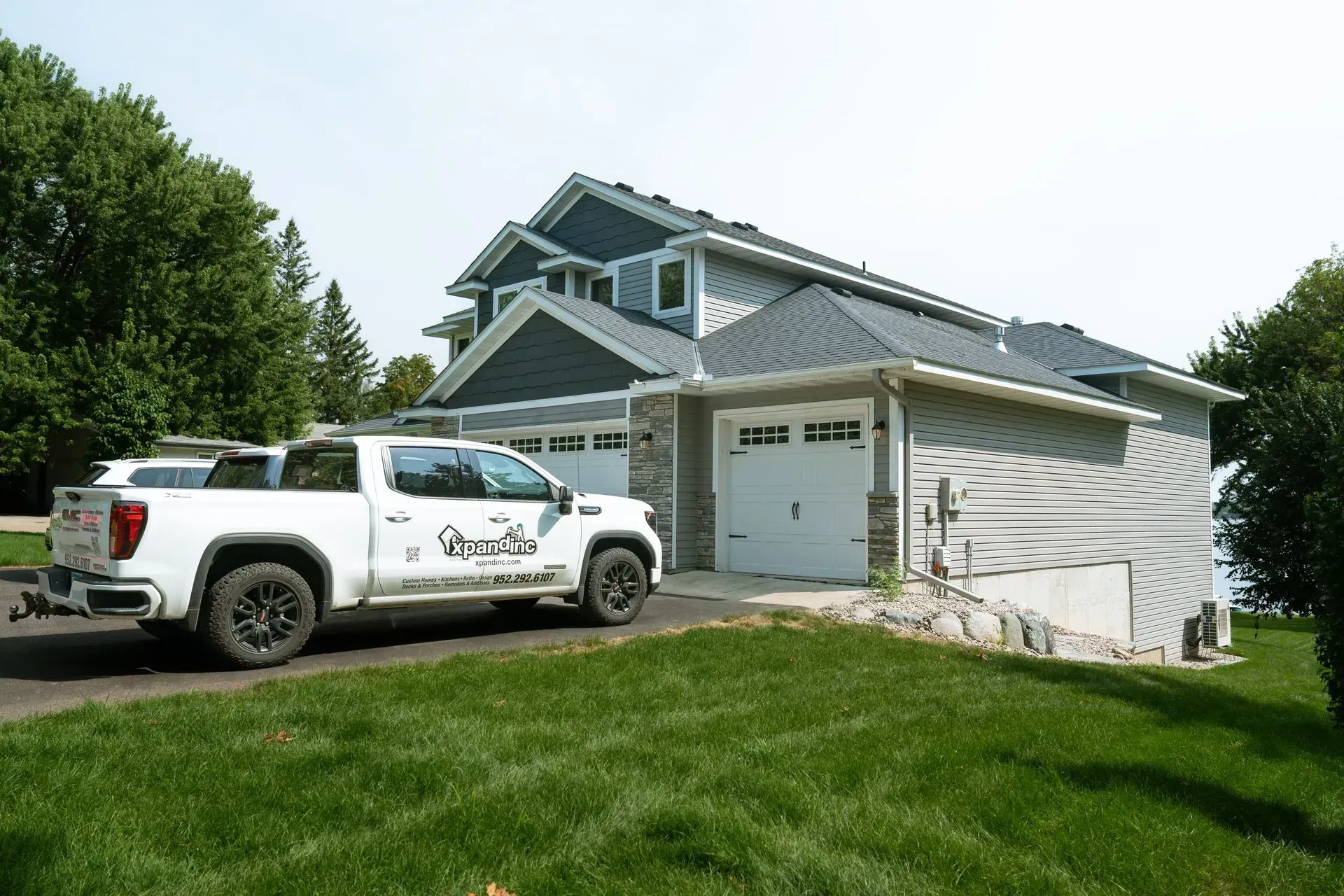 White pickup truck parked in front of a gray house with a two-car garage. Green grass and trees surround.