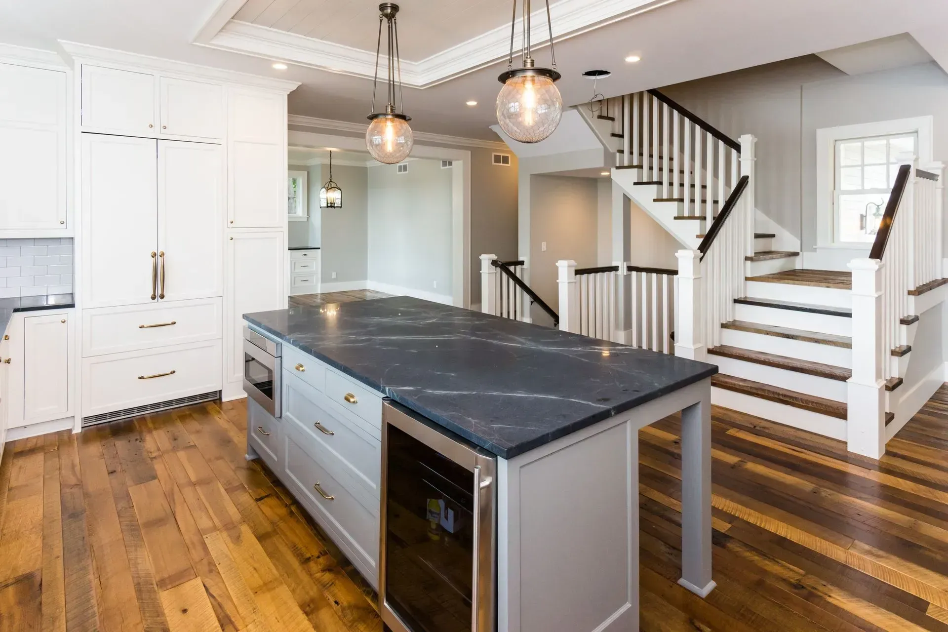 Kitchen with gray island, dark countertops, wine cooler, and hardwood floors. Stairs in the background.