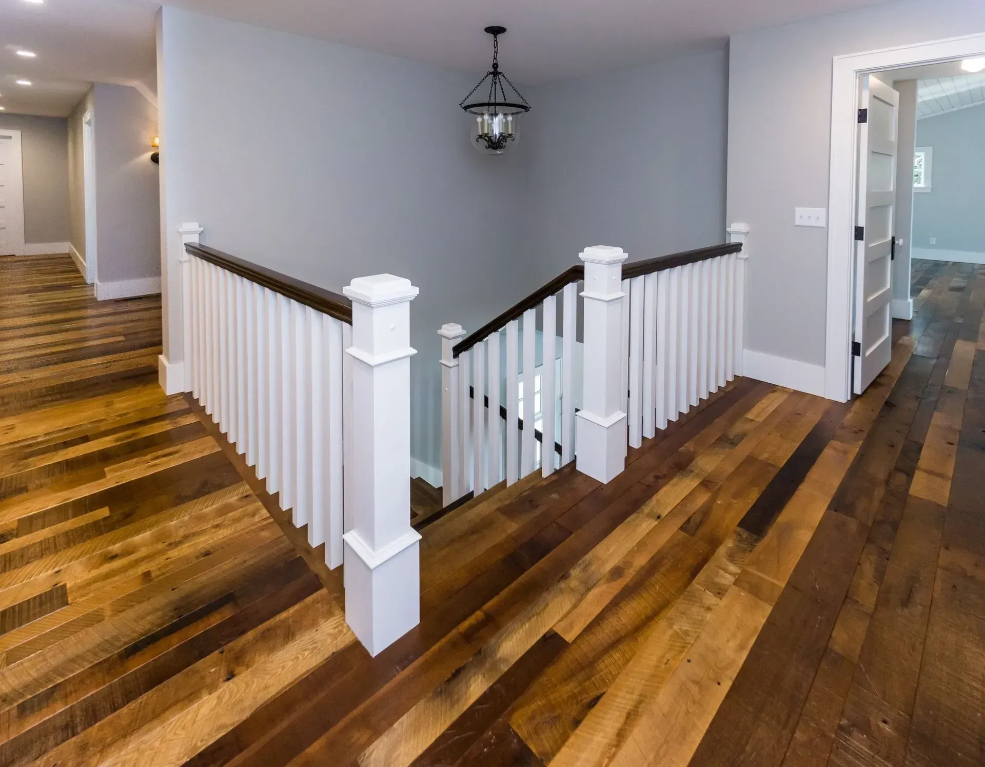 Hardwood floored hallway with white railing around a staircase. Gray walls, black overhead light.