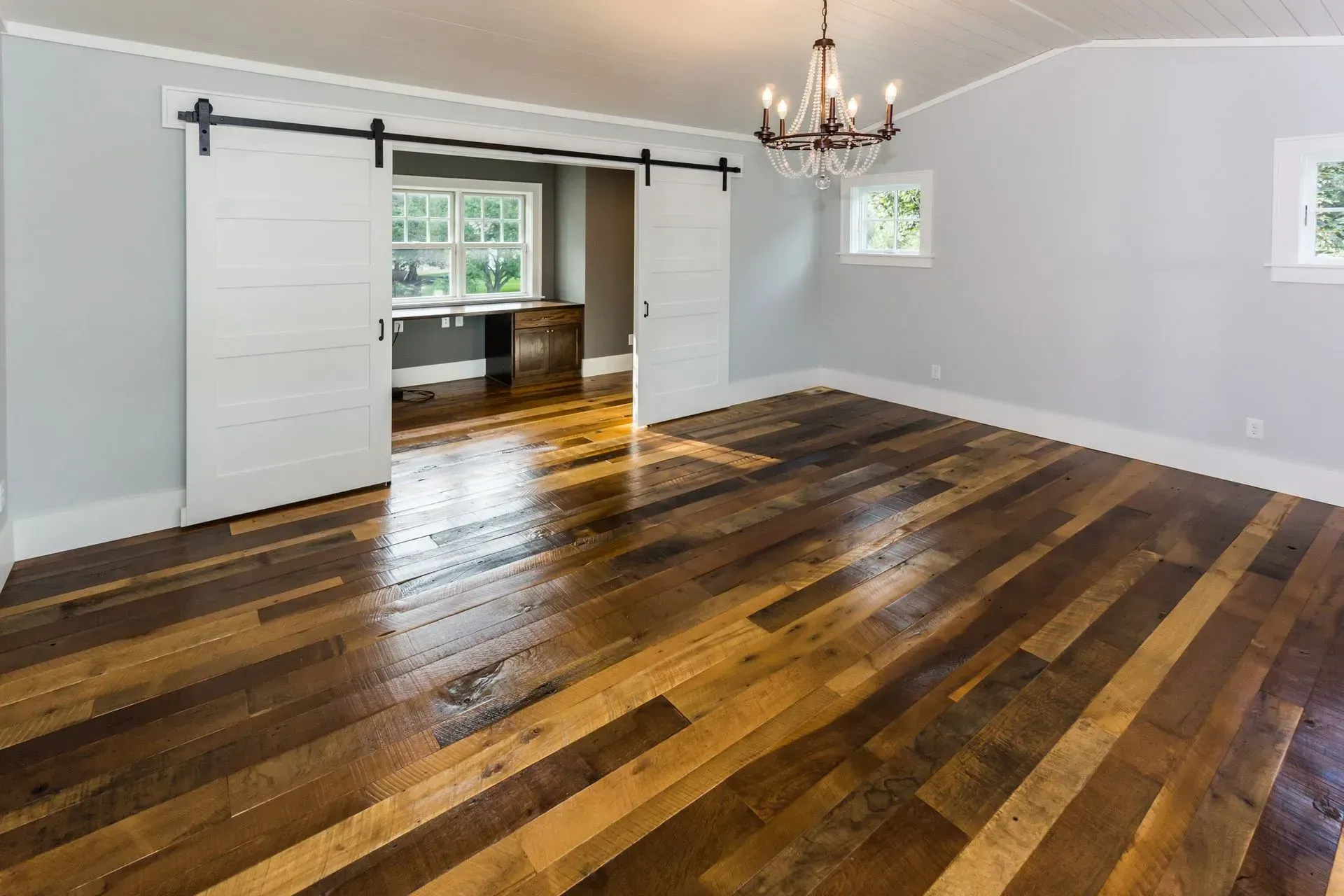 Spacious room with wood floor, gray walls, white sliding doors, and a chandelier; opens to a room with a desk.