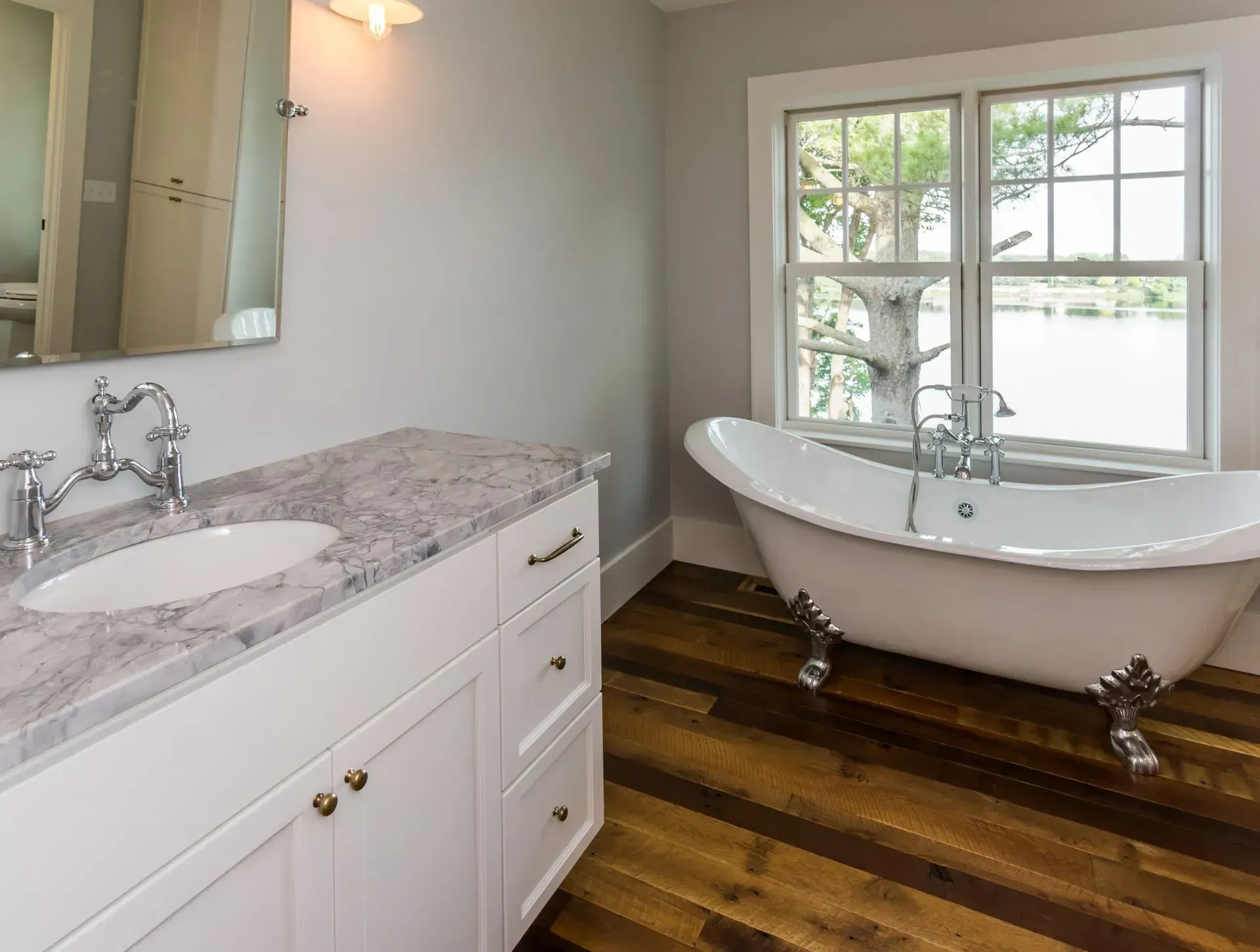 Bathroom with white clawfoot tub, wood floor, marble vanity, and window overlooking water.