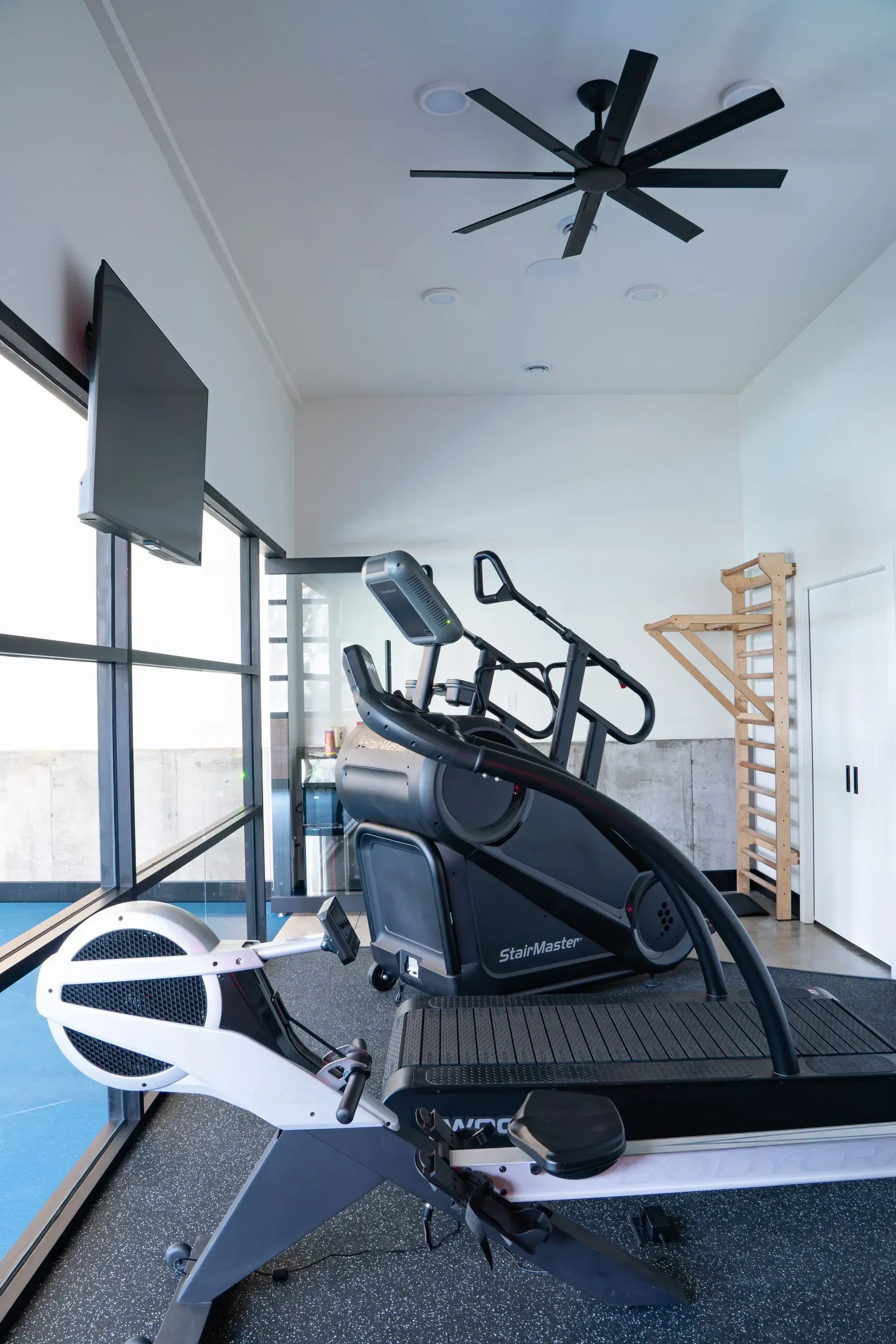 Gym interior with exercise equipment, including a treadmill and rowing machine, and a ceiling fan.
