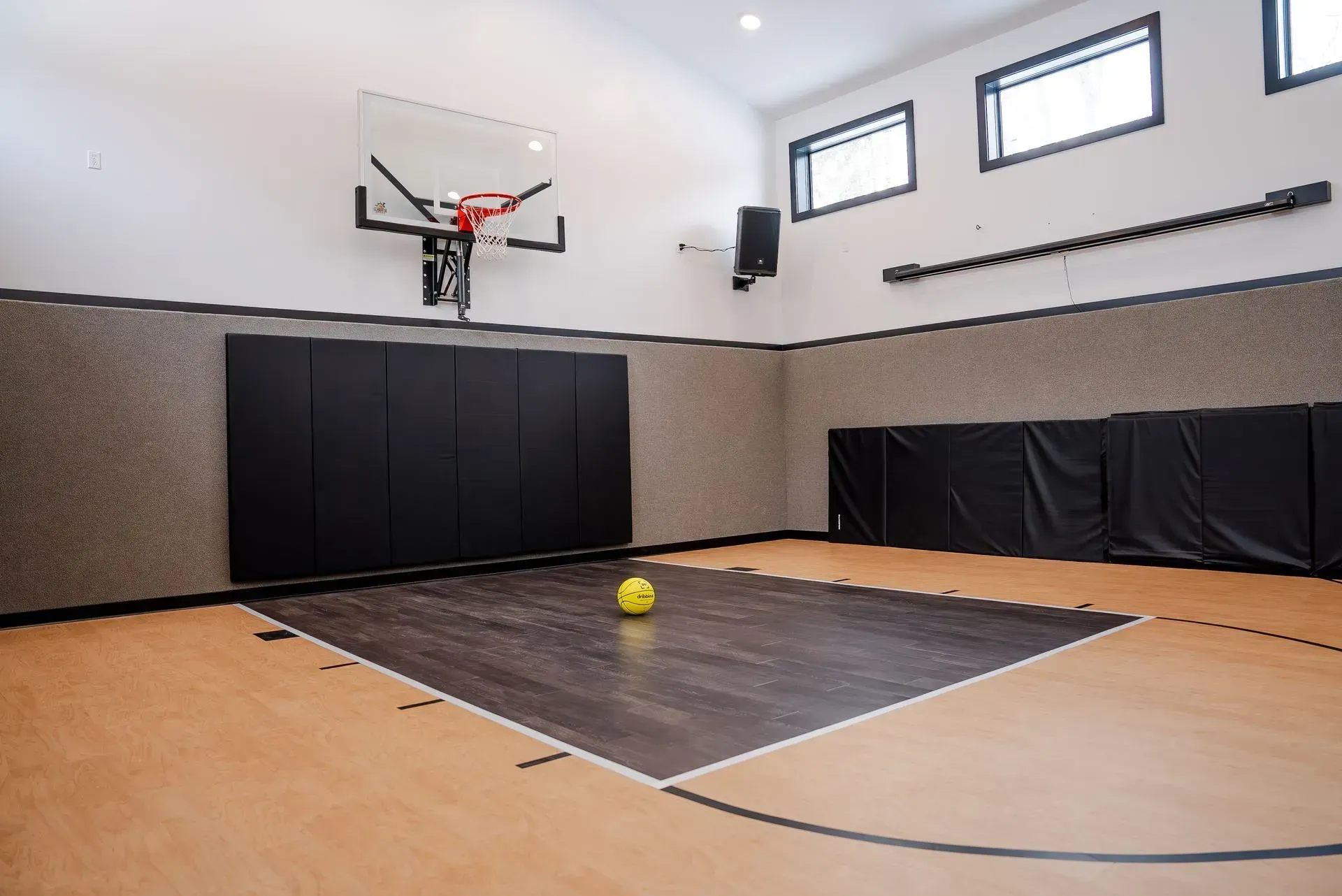 Basketball court with hoop, ball on floor, black padded walls. Light wood and dark gray flooring.