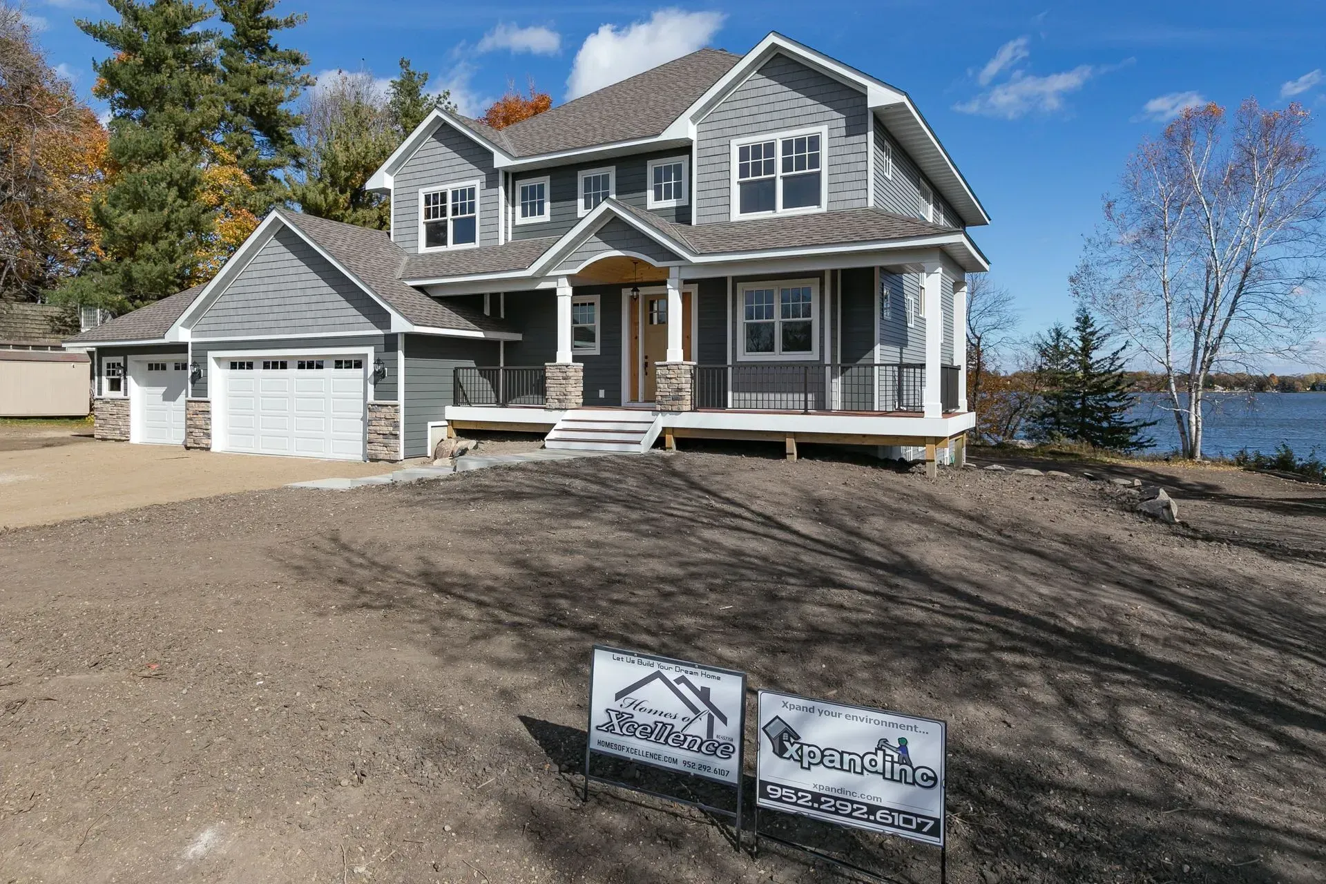 Two-story gray house with white trim, garage, and porch. Located near a body of water with signs in front.