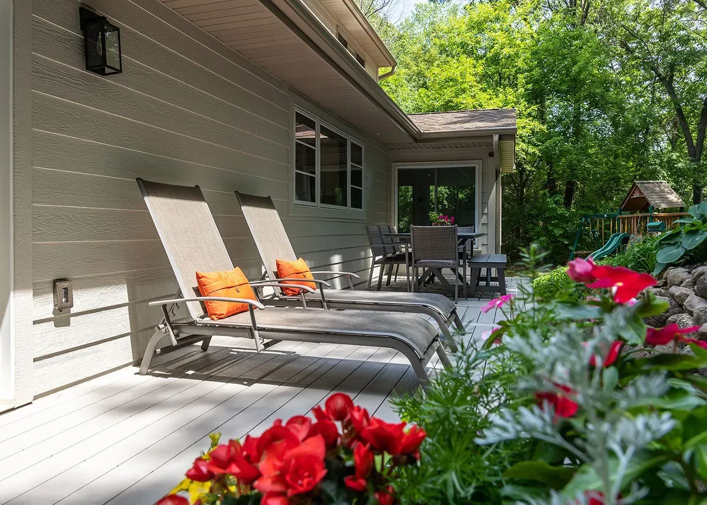 Deck with lounge chairs and table, surrounded by flowers, next to a house with trees.