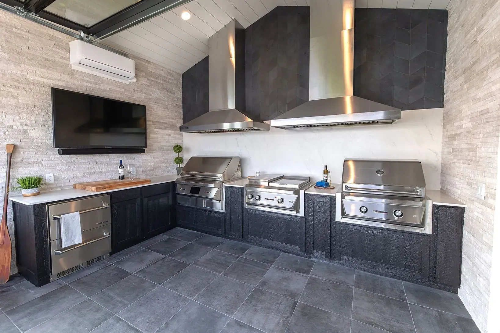Outdoor kitchen with stainless steel grills, range hoods, and a mounted TV.